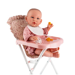 Doll in pink high chair with tray and bib from Asi Dolls Martina Collection, featuring floral fabric and white metal frame.