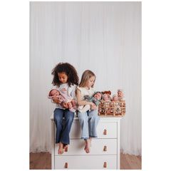 Two children sitting on a dresser holding Asi Dolls, with more dolls in a basket, in a softly lit room.