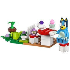 Bluey, a blue dog figure, sits on a red mushroom-shaped stool at a colorful tea party set. The table displays a teapot, cups, and a cupcake, with potted plants and a white picket fence in the background.