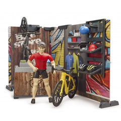 Toy cyclist in a red shirt and helmet stands by a yellow bike in a colorful Bruder bworld bike shop playset.