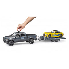 Hand interacting with a gray Bruder RAM Power Wagon toy towing a yellow Roadster on a trailer.