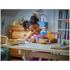 A young girl with curly hair, wearing a purple outfit, smiles while assembling the LEGO Disney Cinderella’s Dress set on a table, featuring a colorful display with Cinderella and a small mouse character.