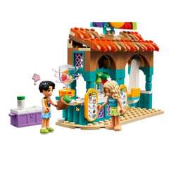 Two mini-dolls, Nova and Niko, stand at the colorful LEGO® Friends Beach Smoothie Stand, surrounded by vibrant smoothie ingredients. A blender whirls on the counter, while the charming beach buggy sits nearby, creating a lively atmosphere for imaginative play.
