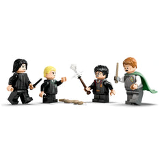 Four LEGO minifigures from the Harry Potter series are positioned in a duel scene. One figure, wearing black robes and holding a wand, appears tense. A second figure, with blonde hair in a green outfit, gestures with an arm raised. The third figure, dressed in a red and gold robe, wields a wand and looks surprised. The last figure, in a green cape, has a worried expression while holding a wand. A flexible brown element representing a magical object lies on the ground