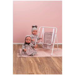 Two dolls in floral outfits sit next to a matching pink and white doll swing on a wooden floor.