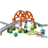 Colorful LEGO® DUPLO® Train Bridge set featuring an elevated orange bridge with green pillars, curved tracks, and a small flower. A figurine stands by a signaling post.
