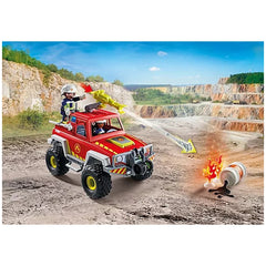 red PLAYMOBIL firefighting truck with two helmeted figures atop, against a rocky cliff and clear sky backdrop.