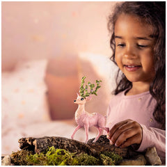 A young girl in a pink top smiles at a small pink toy deer resting on a moss-covered log in a cozy bedroom setting.