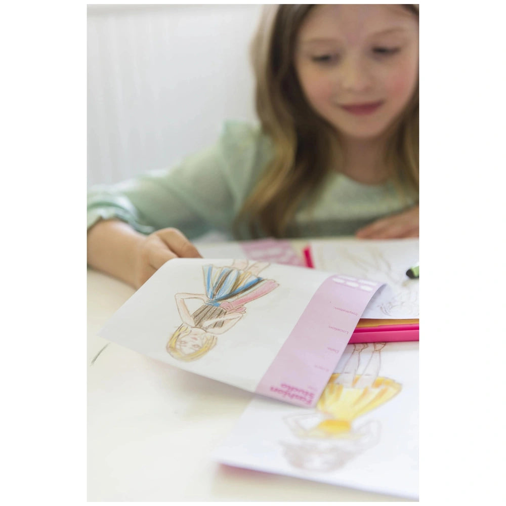 KidzMaker fashion studio kit showcasing hands holding a white printed design paper with a pencil on the table.