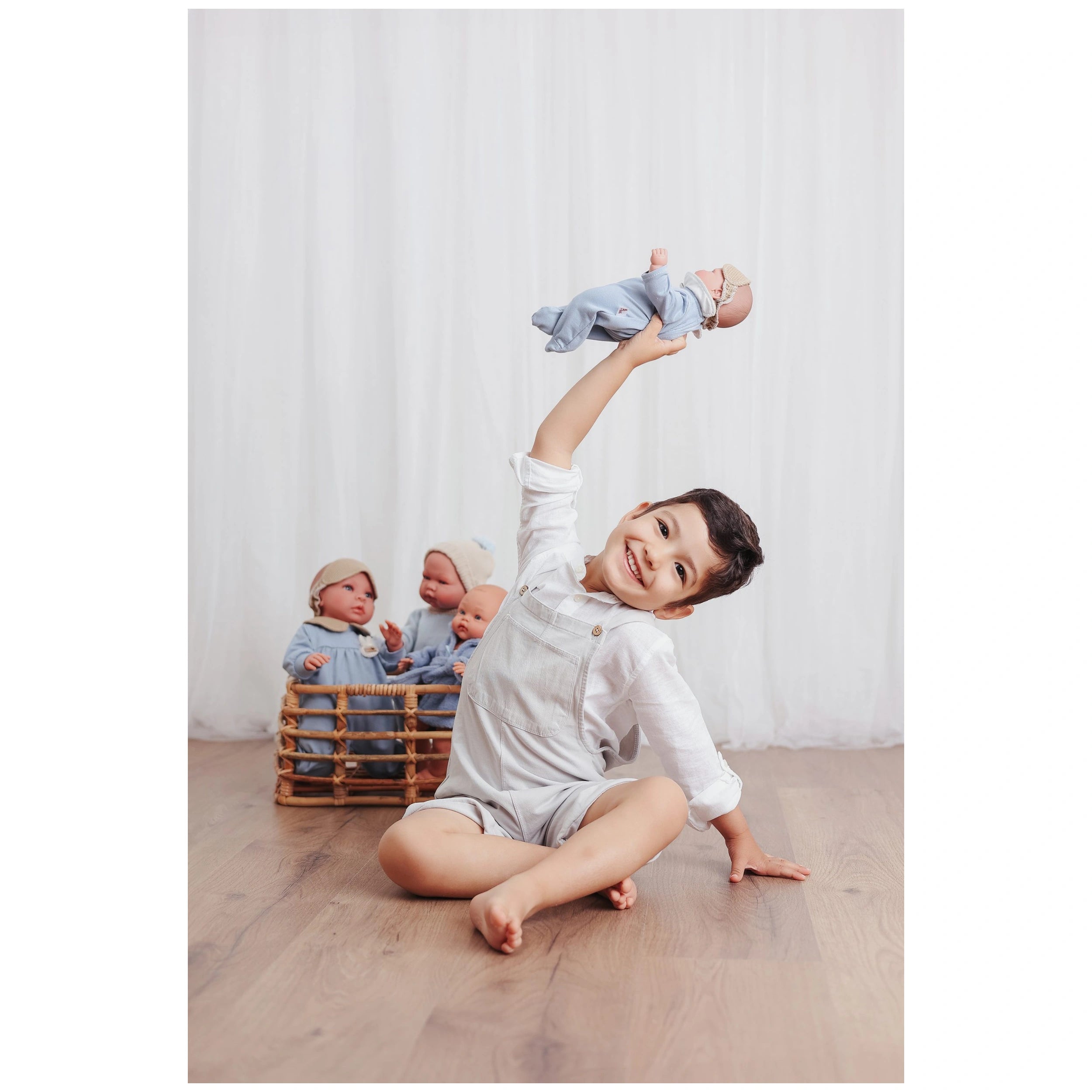 Child in white overalls joyfully lifts ASI doll in blue pajamas, with more dolls in a wicker basket.