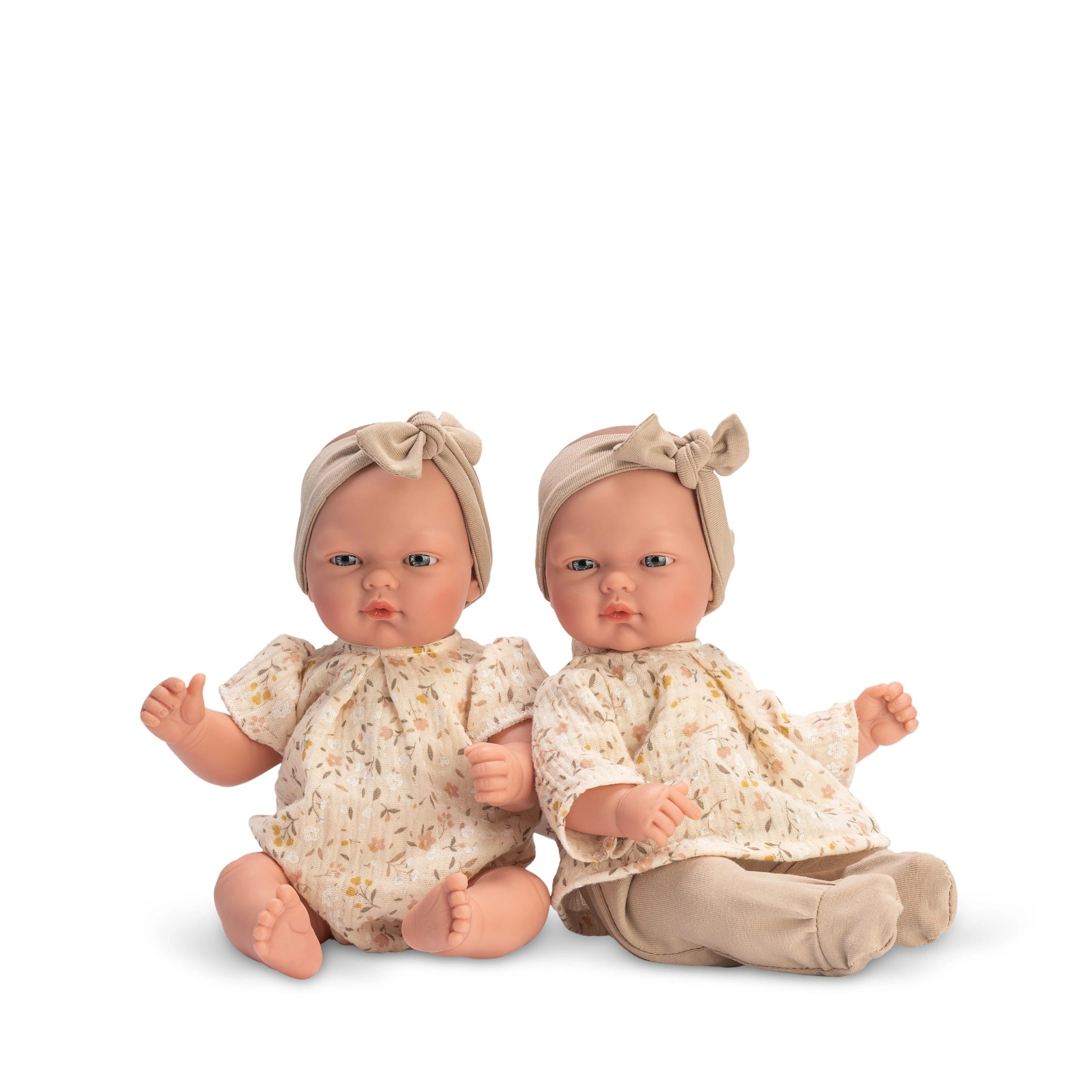 Two ASI Dolls wearing floral rompers and matching headbands sit side by side, showcasing warm expressions and articulated limbs.