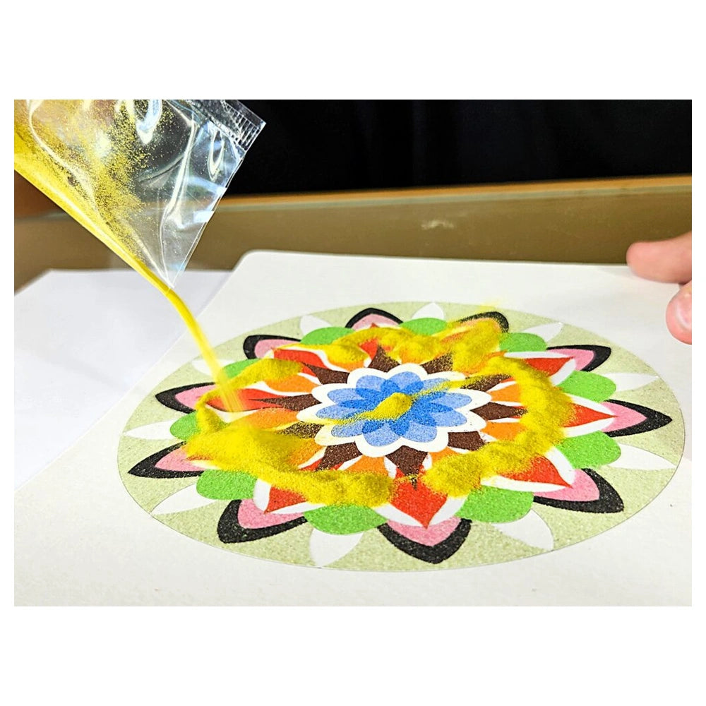 Sand Art Mandala kit featuring a person's hand holding a bag of yellow powder above a piece of cardstock in a blurred indoor 