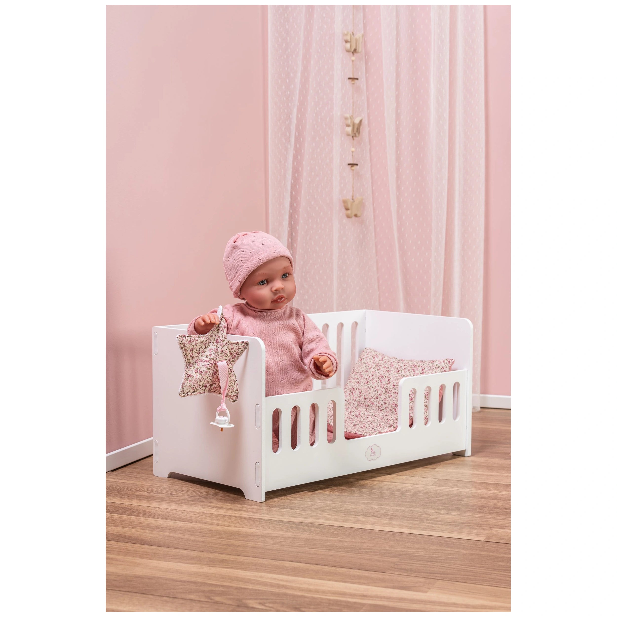 Doll in a white wooden cradle with floral bedding, set in a pink-themed room with sheer curtains.
