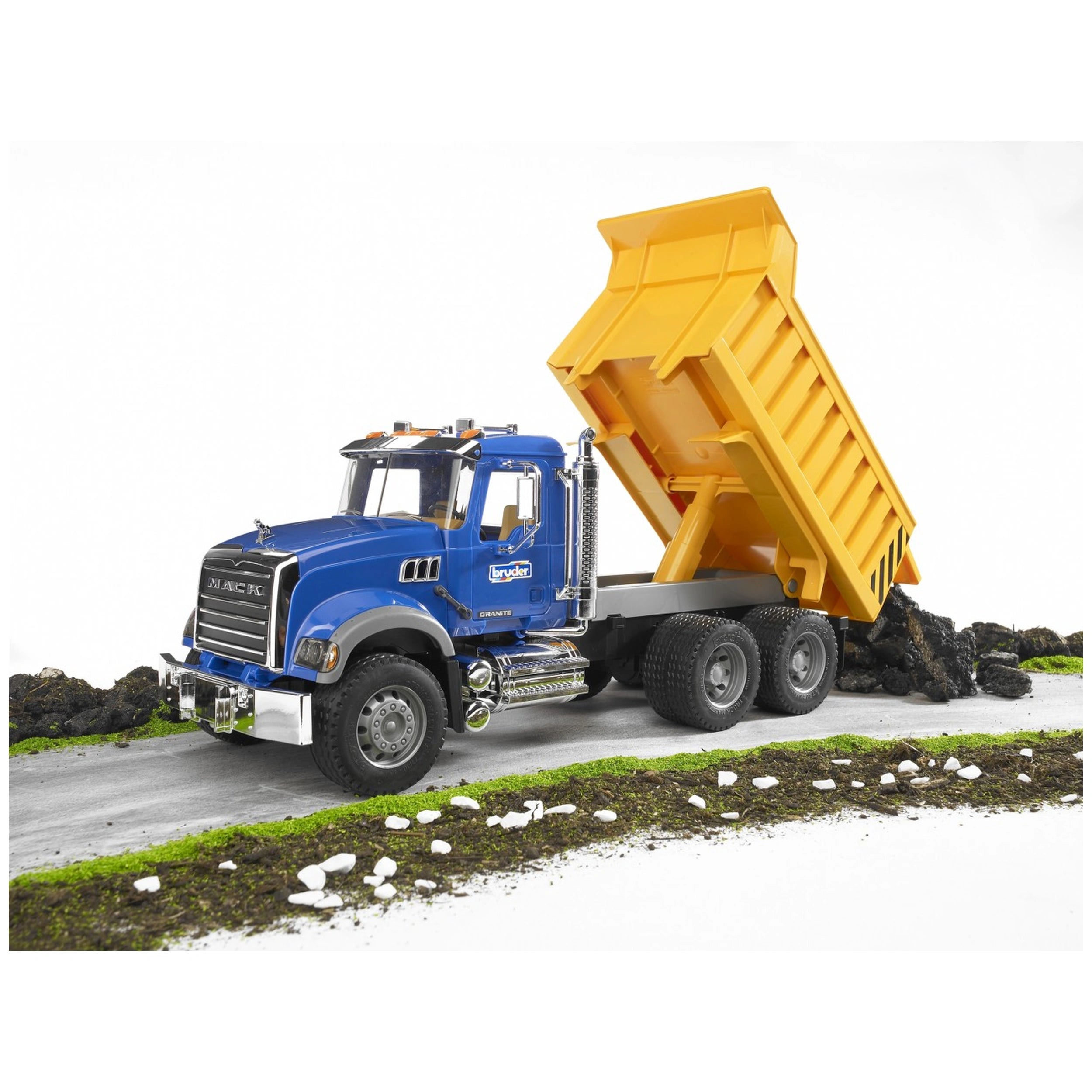 Blue Bruder MACK Granite toy truck with yellow tip-up bed, dumping rocks on a road with green grass.