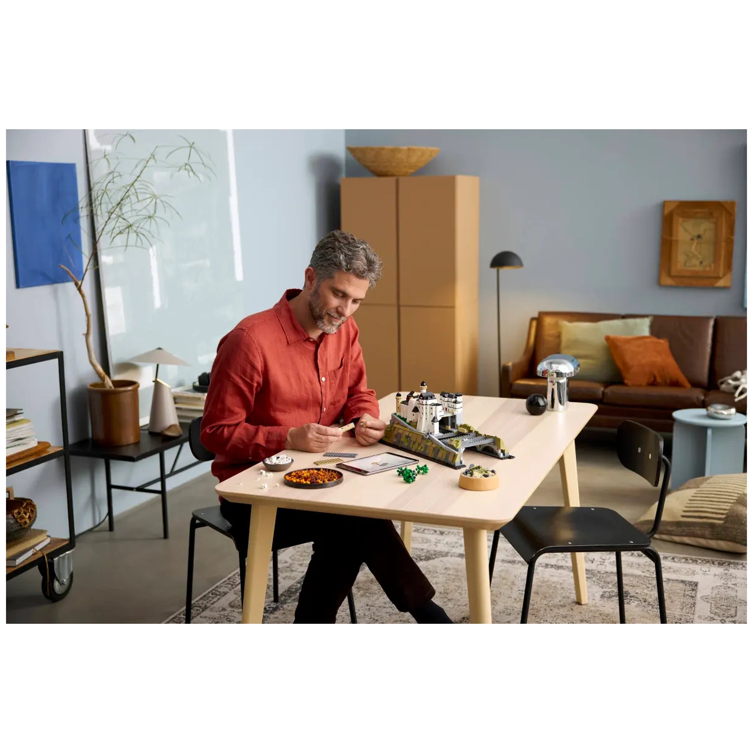 A man in a red shirt sits at a light wooden table, building the LEGO Architecture Neuschwanstein Castle. He smiles while working, surrounded by colorful building pieces, a notepad, and a bowl of snacks. The warm, cozy room features a brown sofa with cushions and a stylish lamp in the background.