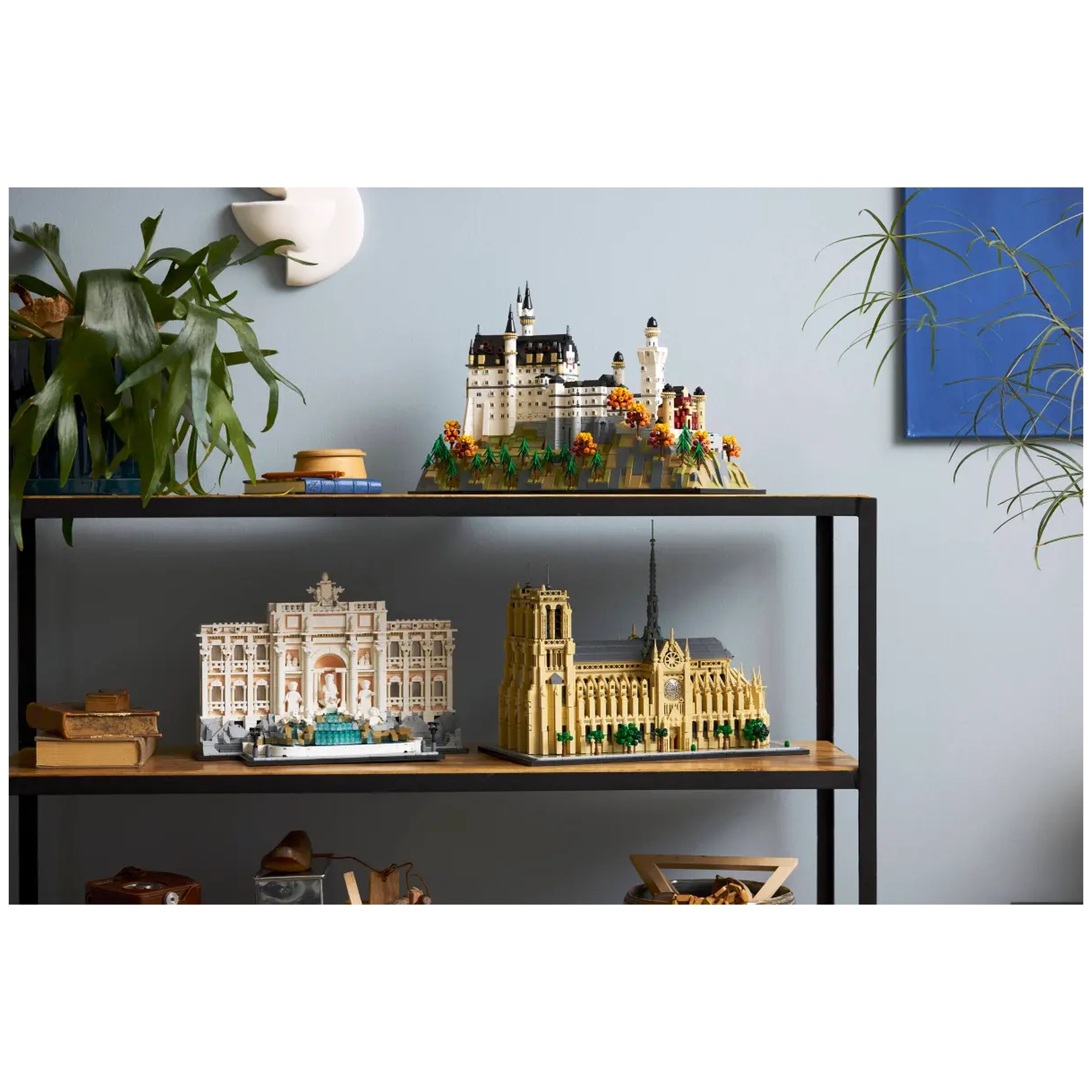 Detailed LEGO architecture models displayed on a black shelf against a light blue wall. The top shelf features Neuschwanstein Castle surrounded by greenery and colorful flowers. The bottom shelf showcases intricate replicas of the Trevi Fountain and a historic building with a clock tower, enhanced by decorative plants and various accessories.