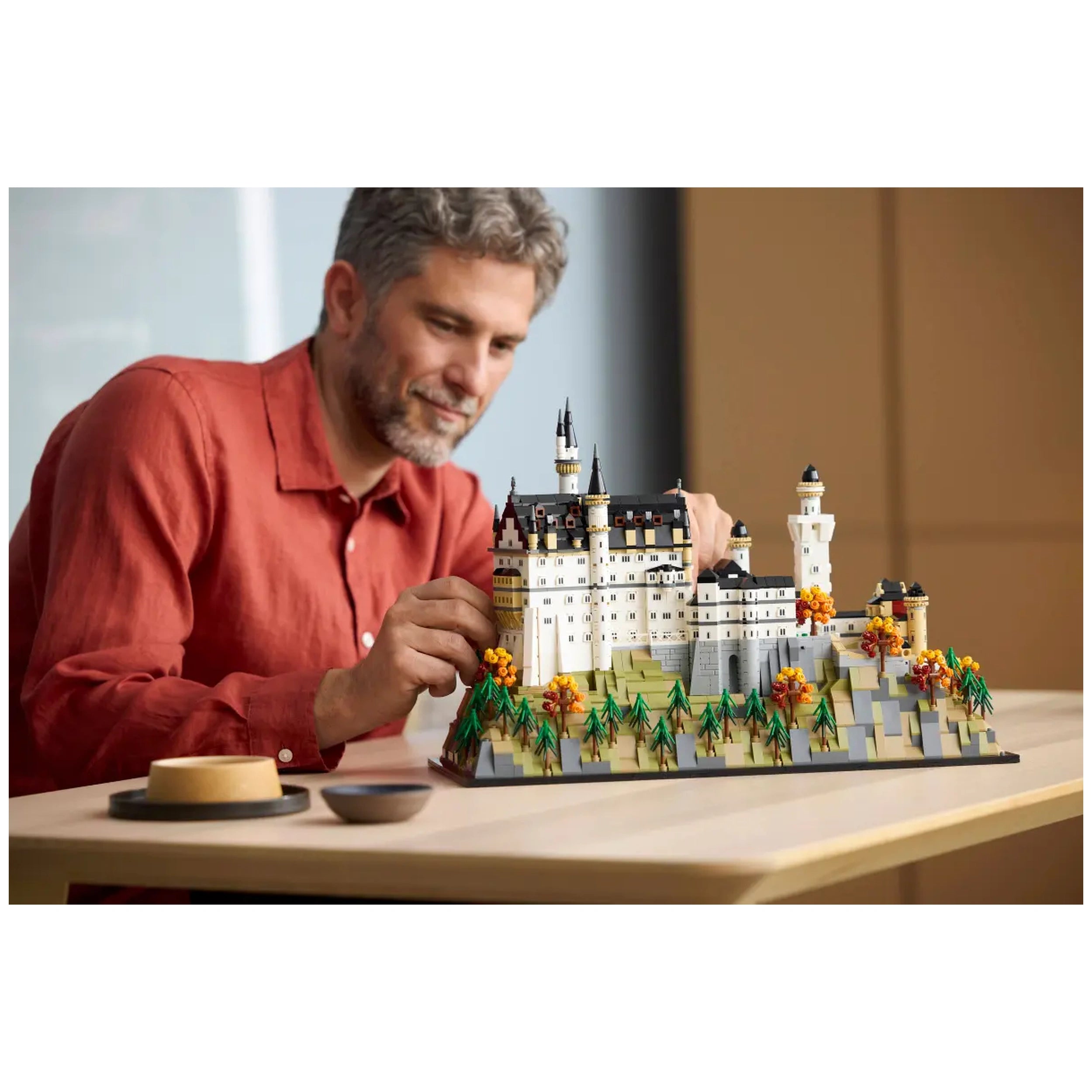 Adult man assembling a detailed LEGO® Architecture model of Neuschwanstein Castle, surrounded by autumn trees on a wooden table.