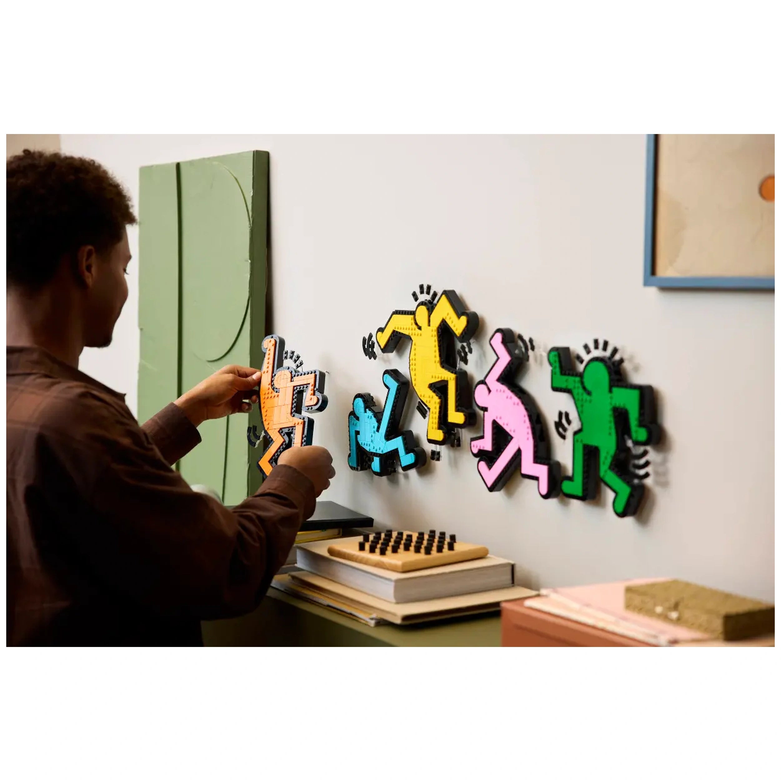 A person wearing a brown shirt carefully attaches a vibrant orange LEGO figure to a wall, surrounded by colorful dancing figures in yellow, green, blue, and pink, inspired by Keith Haring’s art. A wooden shelf below displays books and a chess set, complementing the creative scene.