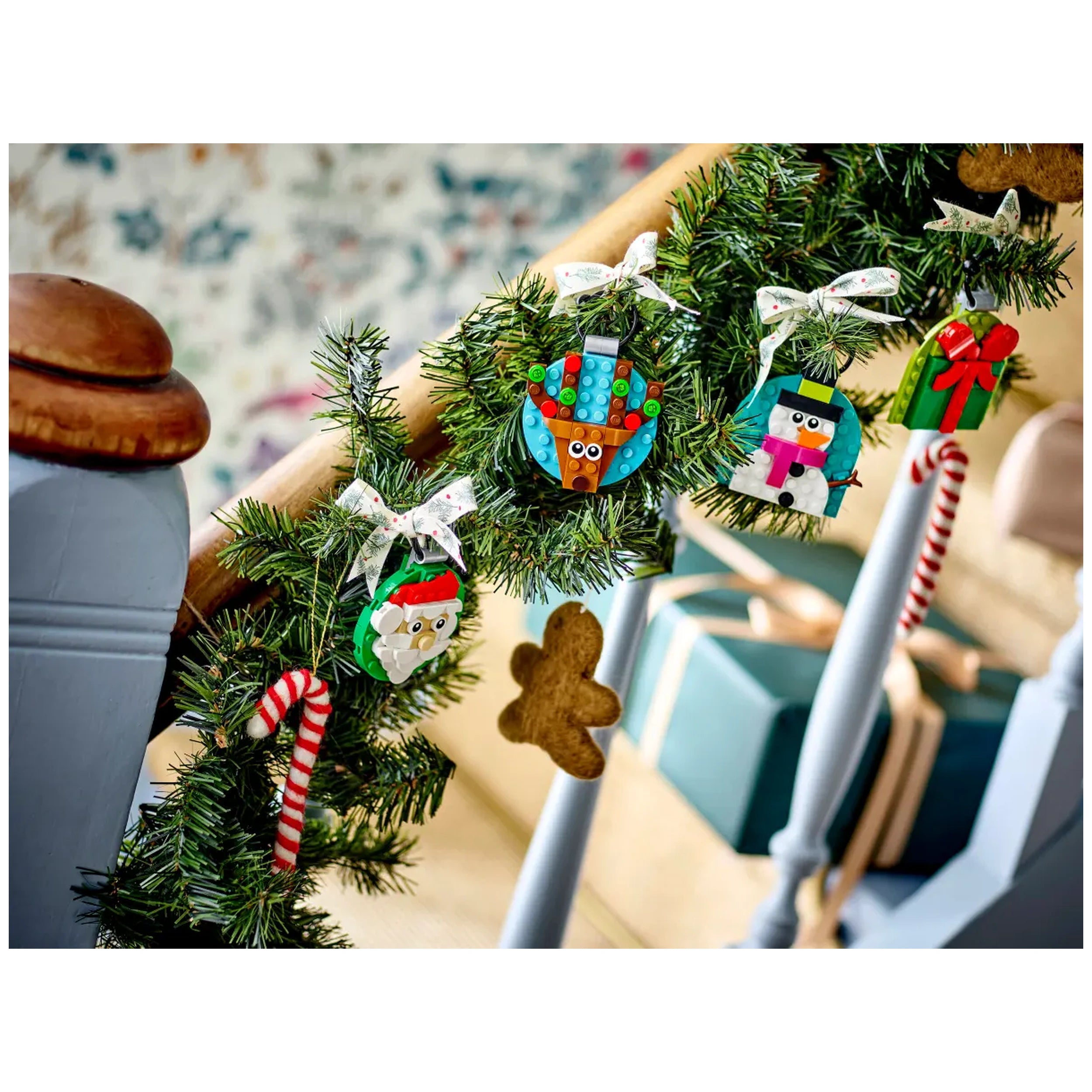 Four colorful LEGO® Christmas ornaments hang from a green garland on a staircase. The ornaments include a jolly Santa, a cheerful snowman, a playful reindeer, and a beautifully wrapped gift, all tied with festive bows. A striped candy cane and a gingerbread man add to the holiday decor.