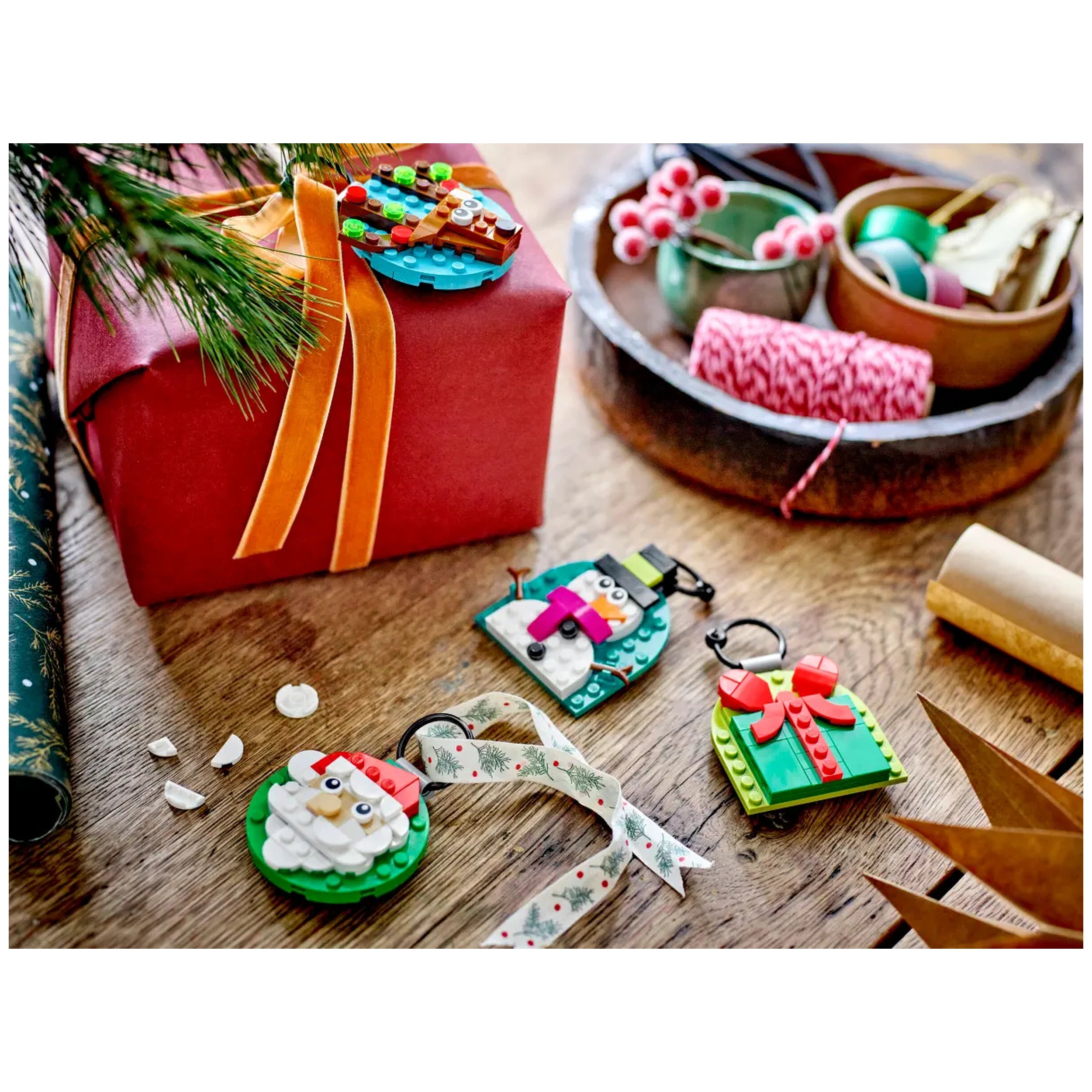 Colorful LEGO® Christmas ornaments, featuring a snowman, Santa, and a gift, beside a wrapped red present on a wooden surface.