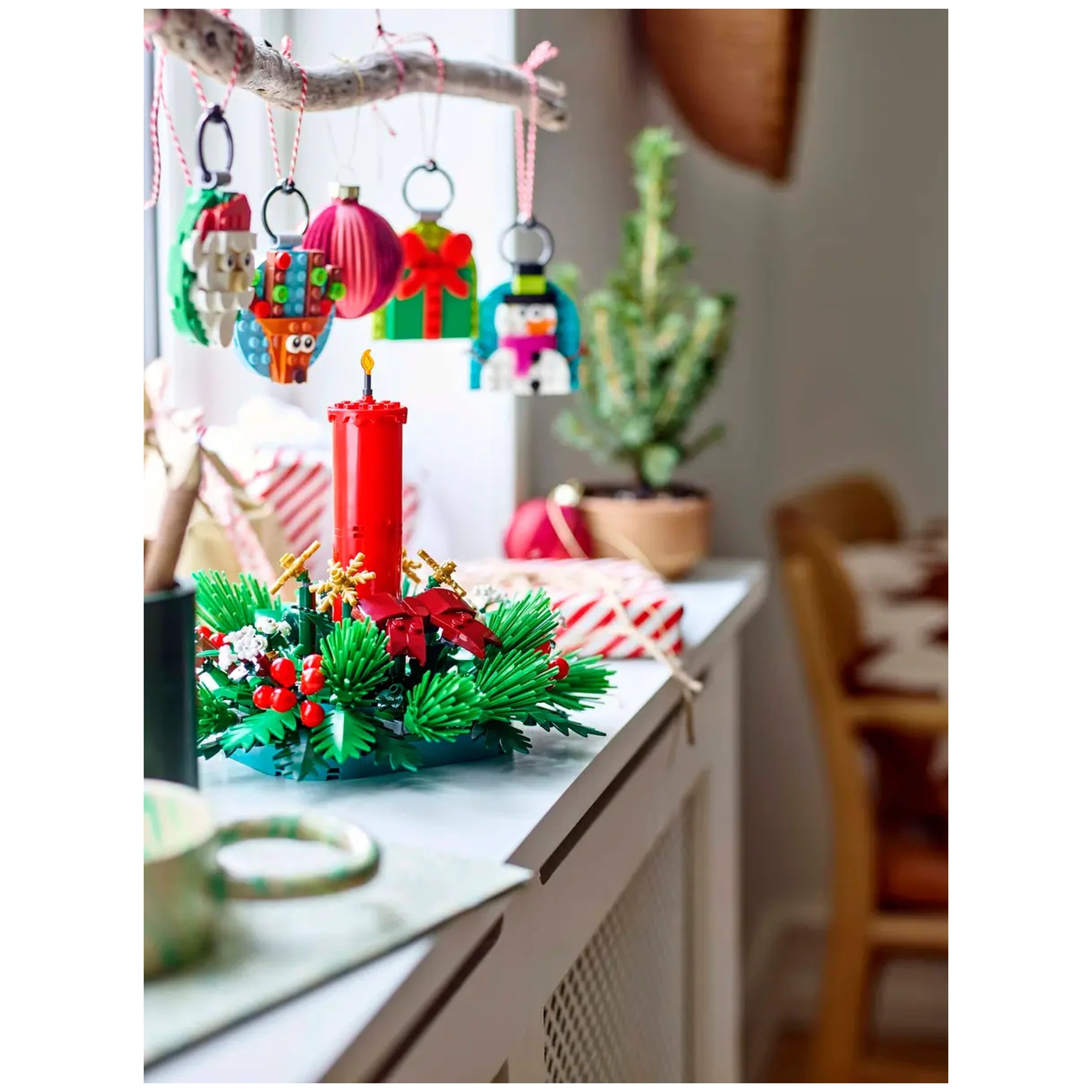 Colorful LEGO Christmas ornaments hang from a twig above a festive centerpiece with a red candle, surrounded by vibrant greenery and berries.