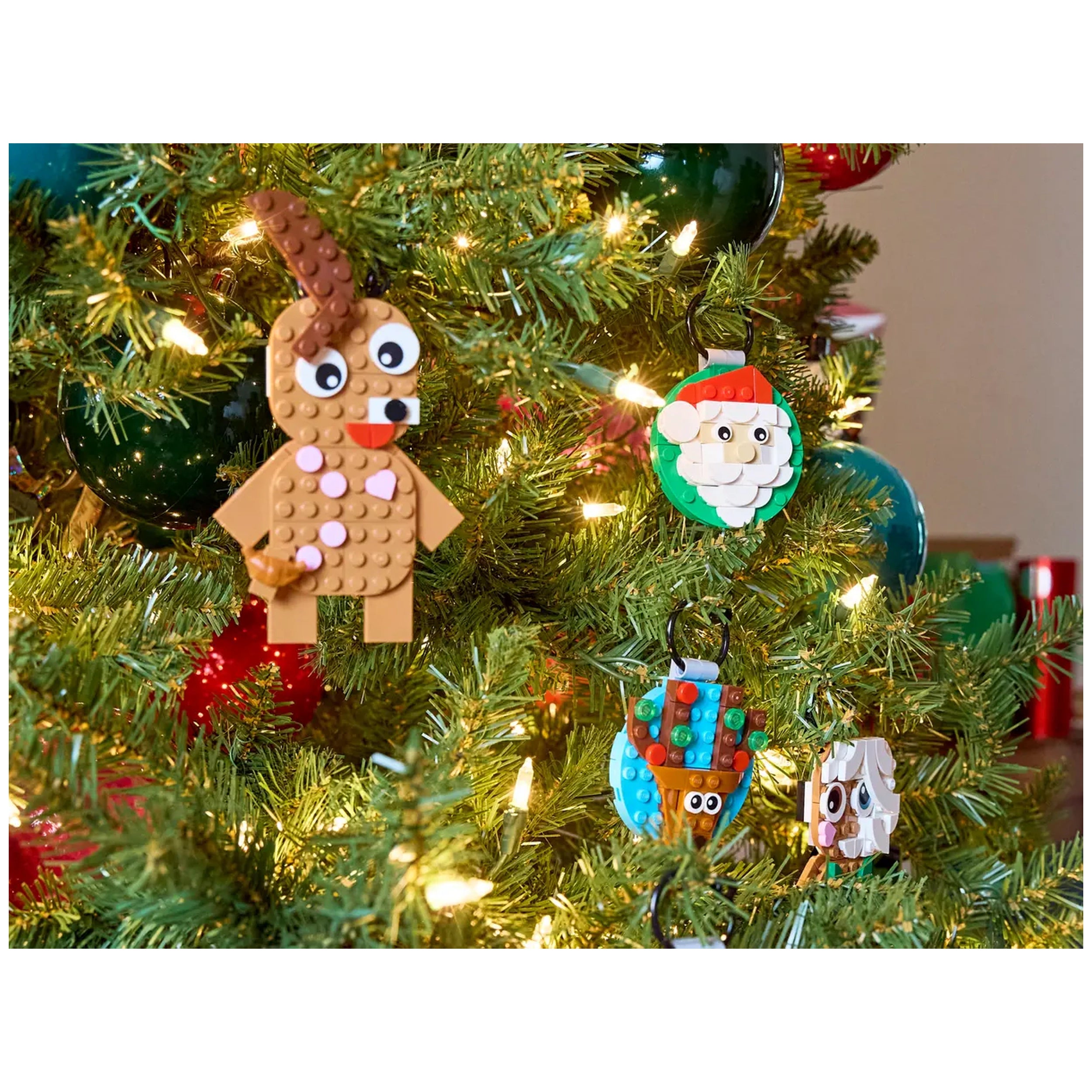 Four colorful LEGO® ornaments hang on a decorated Christmas tree. A gingerbread man with a candy cane, a jolly Santa, a cheerful snowman, and a playful reindeer add festive charm. Twinkling lights enhance the cozy holiday atmosphere.