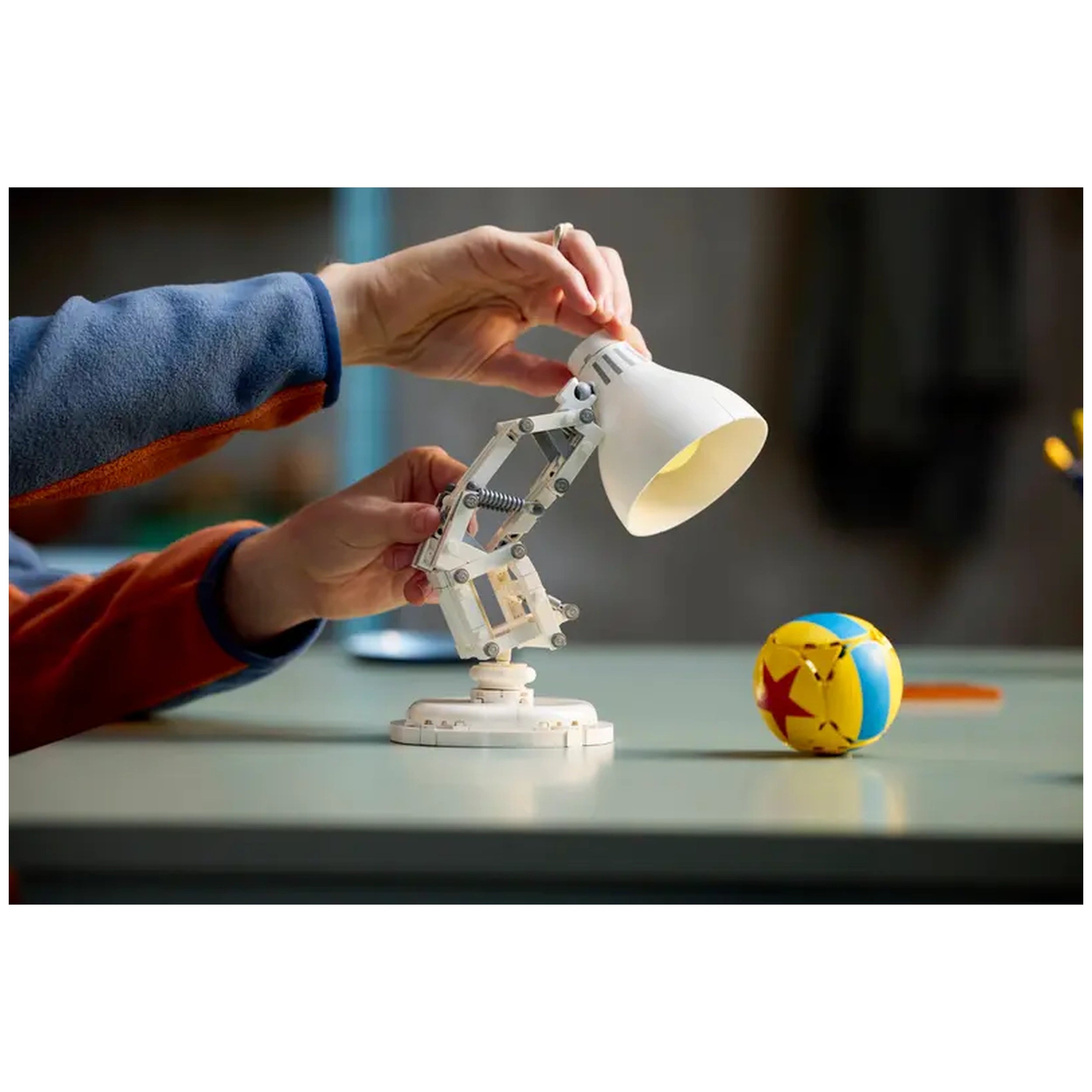 A hand adjusts the lamp of a LEGO Luxo Jr. model, poised next to a colorful ball featuring a star design, on a light-colored surface.