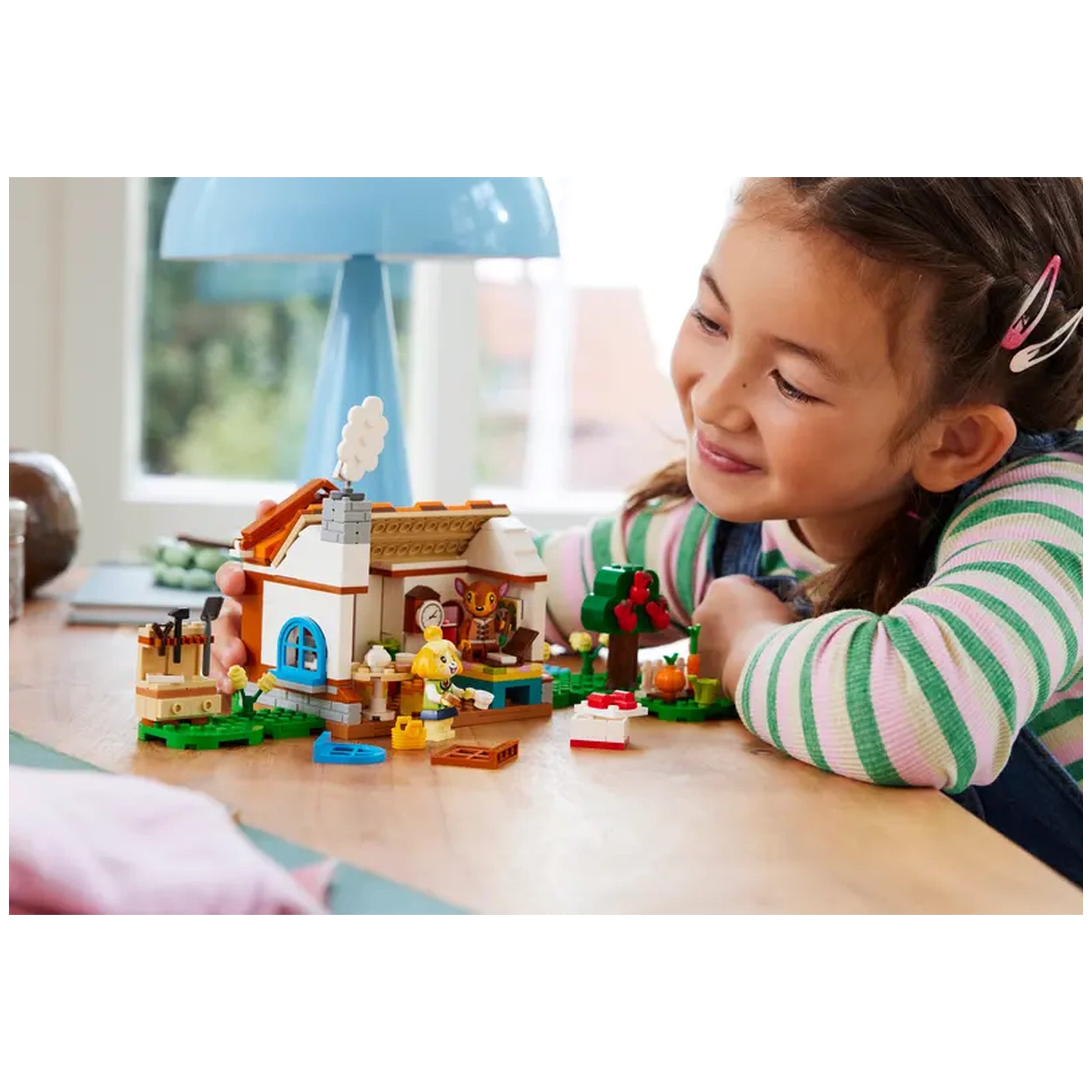 A young girl smiles while playing with the LEGO® Animal Crossing™ Isabelle’s House Visit set on a wooden table, surrounded by colorful building pieces. The charming house features a detailed interior, and a green tree is nearby. A blue lamp and a cozy atmosphere create a playful ambiance.