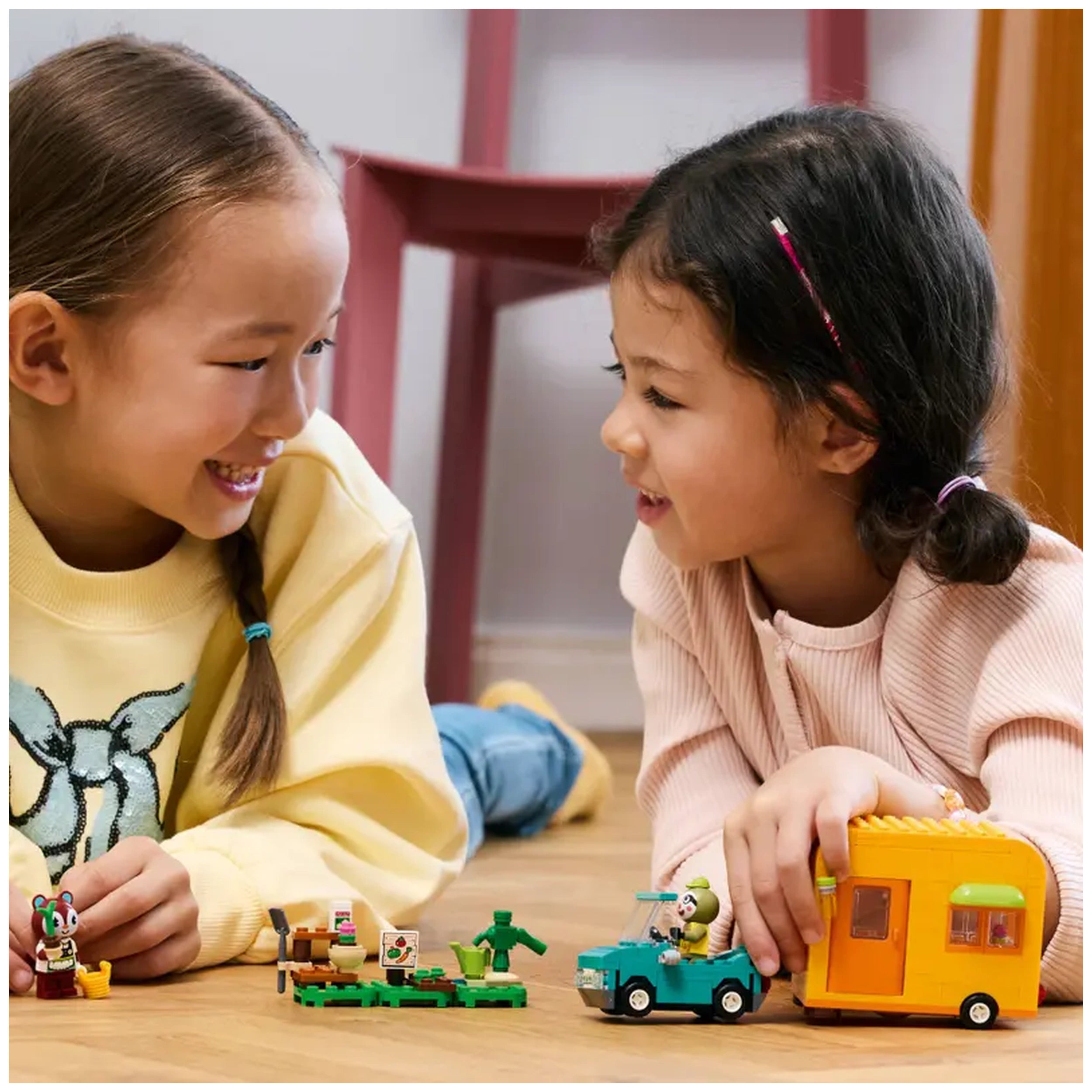 Two smiling girls play on the floor with the LEGO® Animal Crossing™ set, featuring mini figures, a green car, and a colorful caravan.