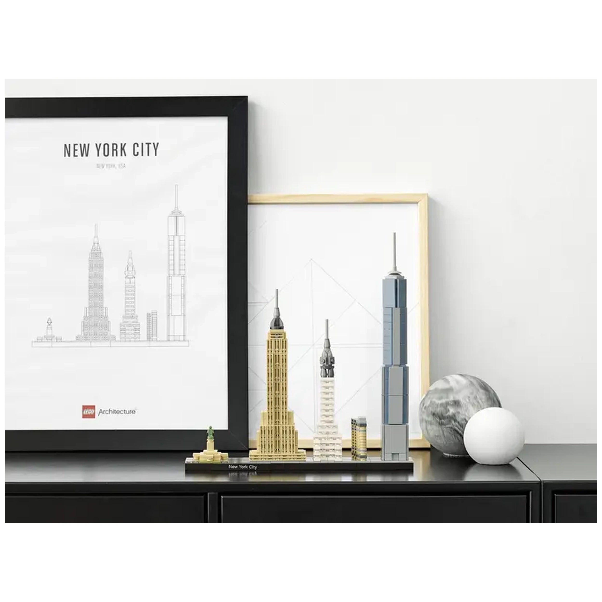 Lego Architecture New York City set displays iconic landmarks including the Empire State Building and One World Trade Center on a black shelf, accompanied by framed skyline sketches and decorative orbs.