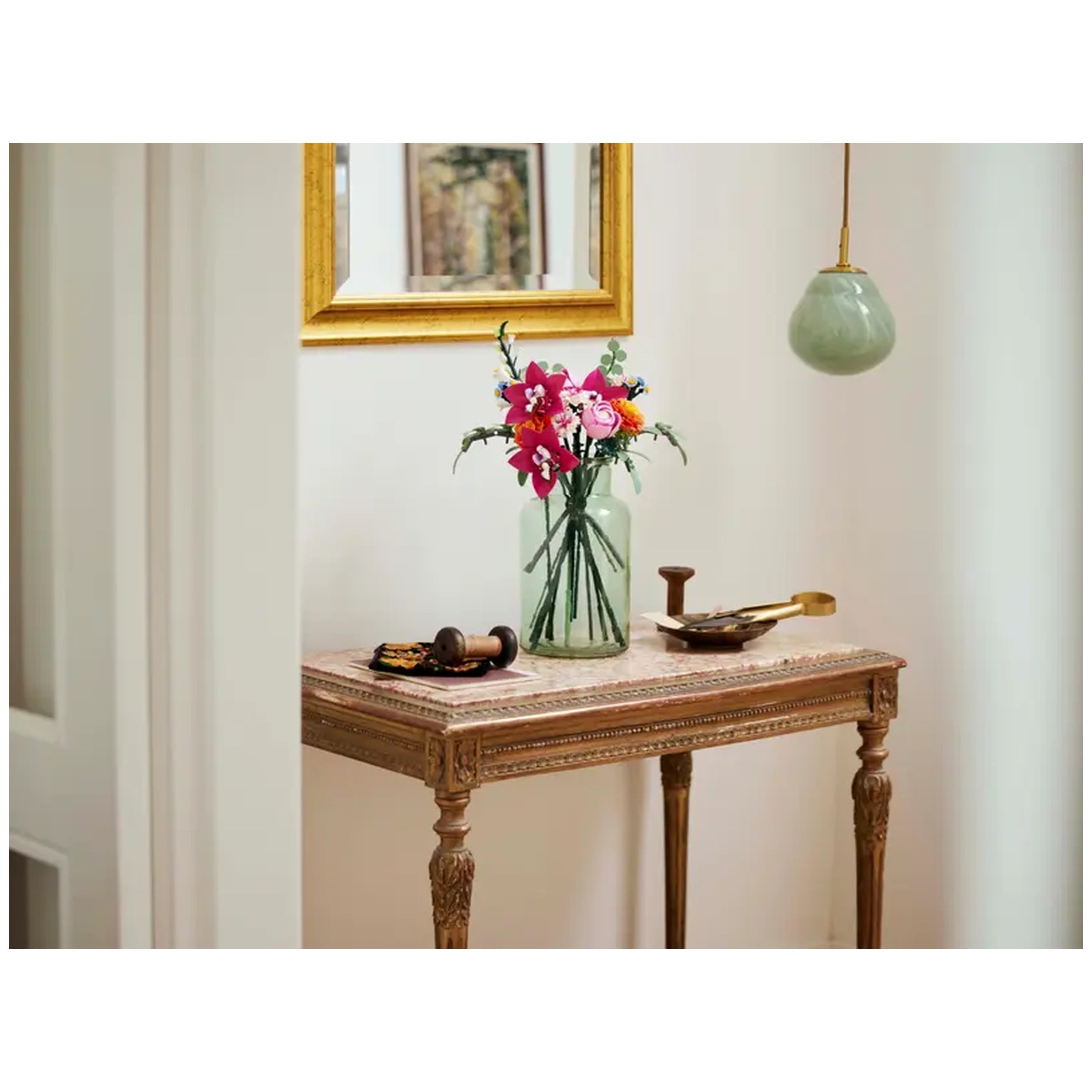 A beautifully arranged Pretty Pink Flower Bouquet in a clear vase, featuring colorful stems of flowers, sits on a vintage wooden table. A decorative bowl and a wooden spool accompany the bouquet, while a soft green pendant light hangs in the background, enhancing the elegant decor.