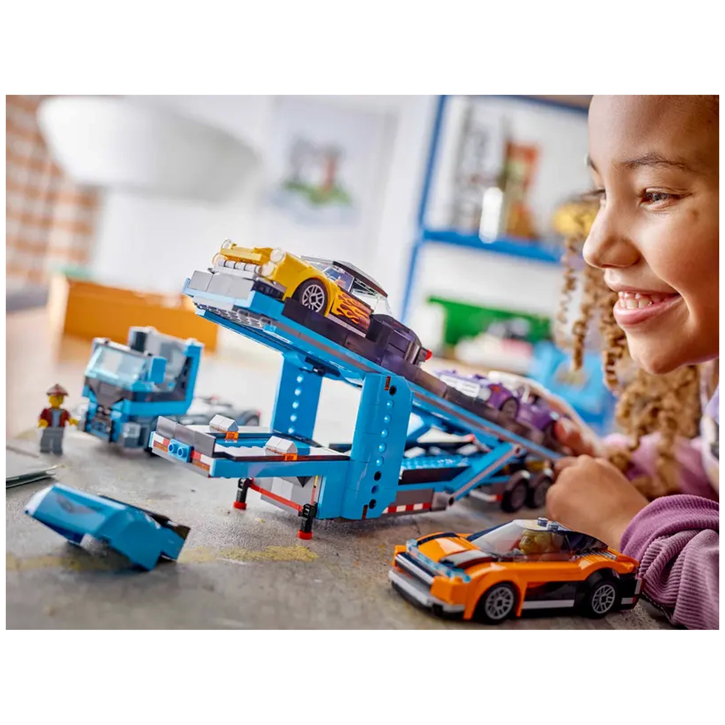 A smiling child with curly hair plays with a colorful LEGO® City Car Transporter, loaded with a yellow sports car and a purple vehicle on its ramps. The blue truck is on a textured surface with a few scattered toy cars nearby, showcasing a fun and imaginative play scene.