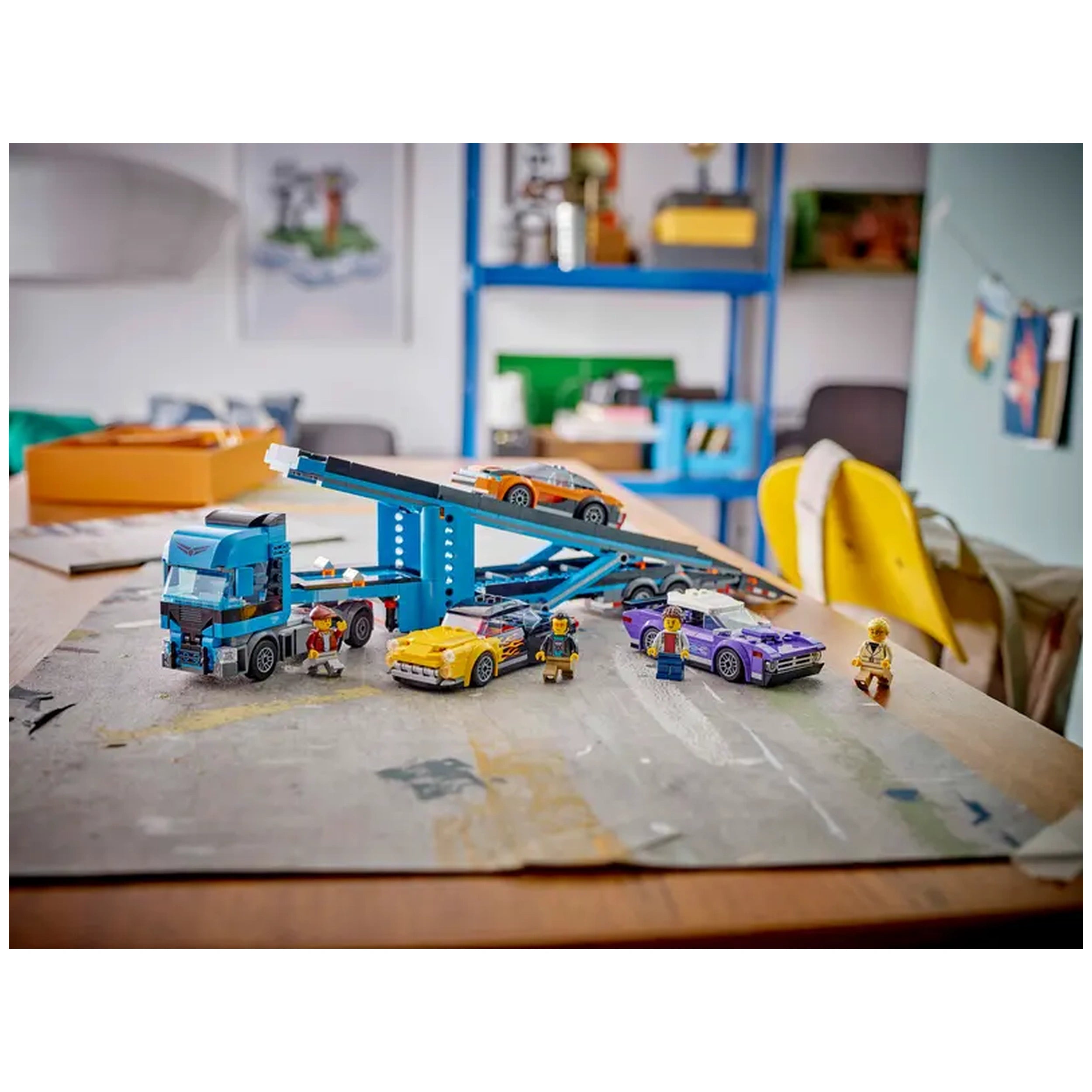 Blue LEGO® Car Transporter truck with a tilted driver’s cab, displaying an engine. A multicolored lineup of sports cars, including a yellow car with flames and a purple car, surrounds it. Two LEGO® figures stand near the vehicles, ready for play. The scene is set on a wooden table in a bright indoor space, with a blue shelf and colorful items in the background.