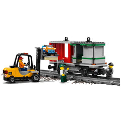 A yellow forklift transports a blue toy car to a gray cargo train car on tracks. Two LEGO mini-figures are present: one in a green hat inspecting the cargo and another in a yellow hard hat. The train car features red accents and is partially open, showcasing an interior with green detailing. Gravel surrounds the tracks, enhancing the construction theme.