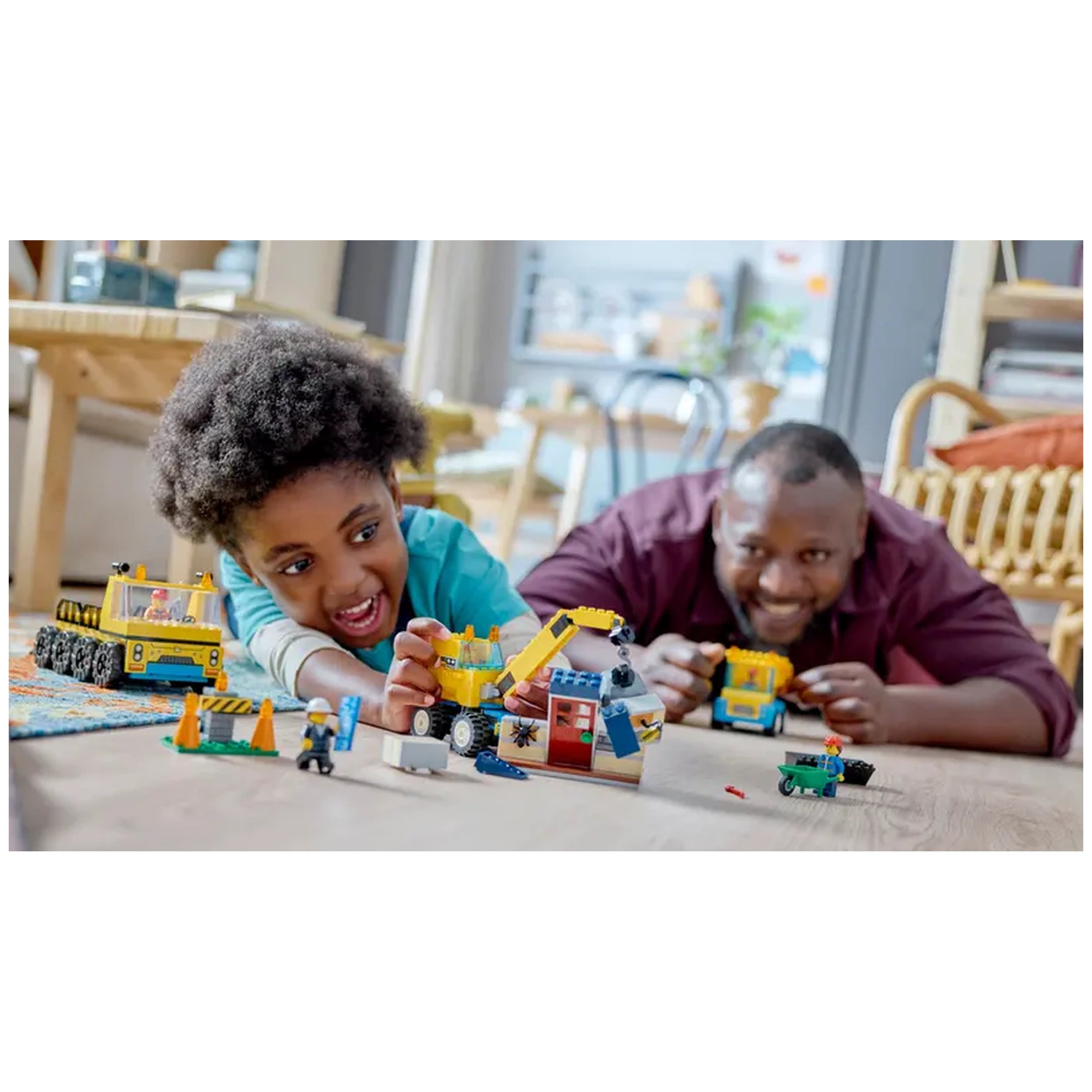 A child with curly hair joyfully plays with LEGO® construction toys, building a wrecking ball crane and nearby vehicles, while an adult smiles, sharing the experience in a cozy, well-lit room.