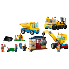 LEGO® City Construction set featuring a yellow dump truck, a wrecking ball crane, and a transporter, along with three worker minifigures and accessories. A derelict house with a red door is included, with colorful construction elements scattered around.