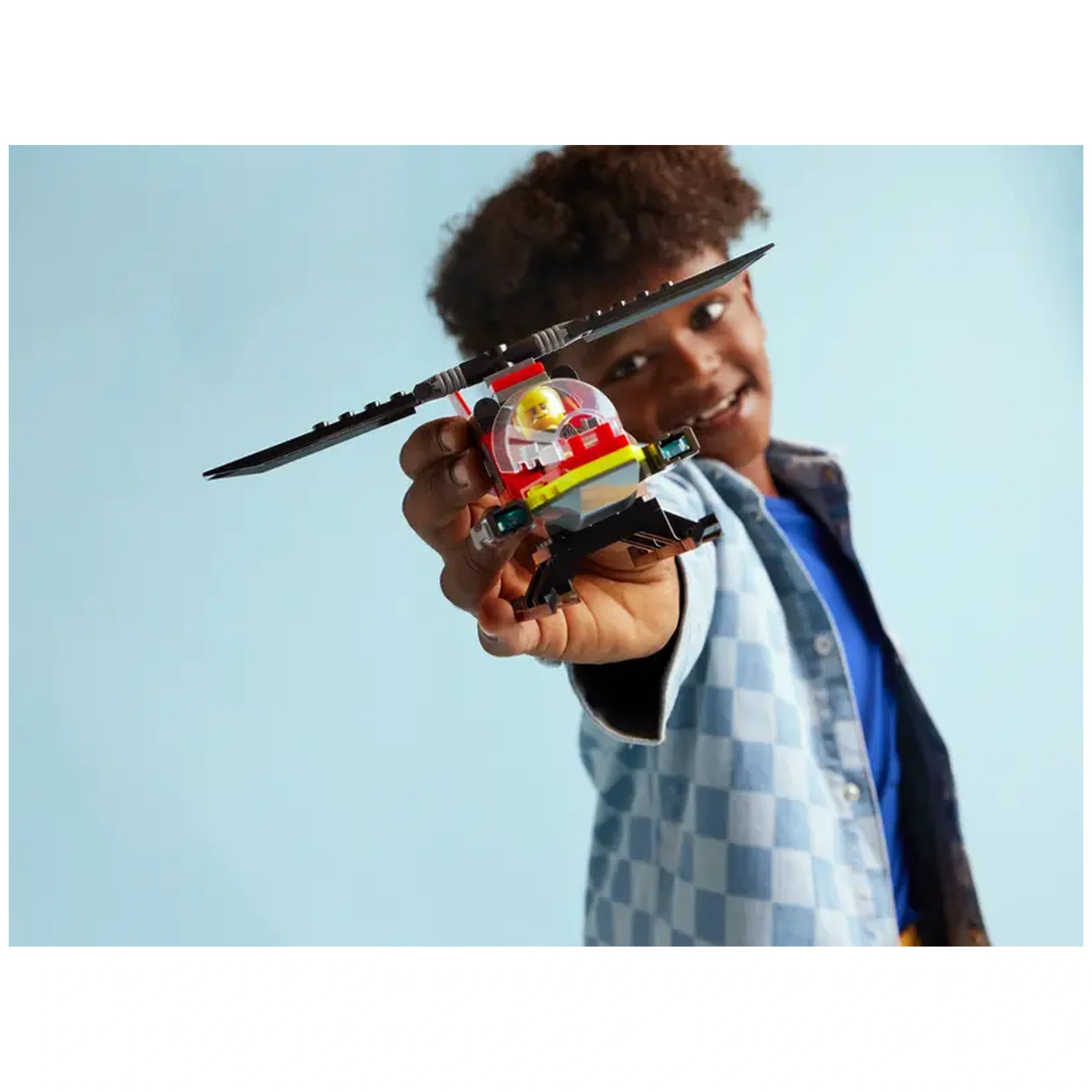 A smiling child holds a colorful LEGO City Fire Rescue Helicopter against a light blue background, showcasing its spinnable rotors and detailed cockpit.