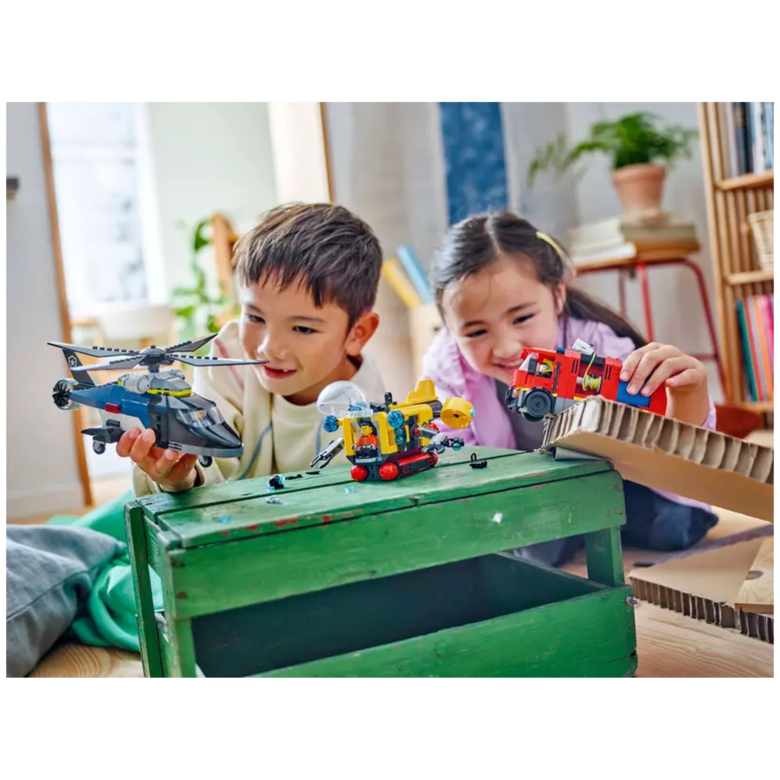 Two children play with LEGO® City toys on a green wooden table. One child holds a helicopter, while the other has a fire truck. A colorful submarine is also displayed on the table. The background features a cozy living space with natural light and bookshelves.