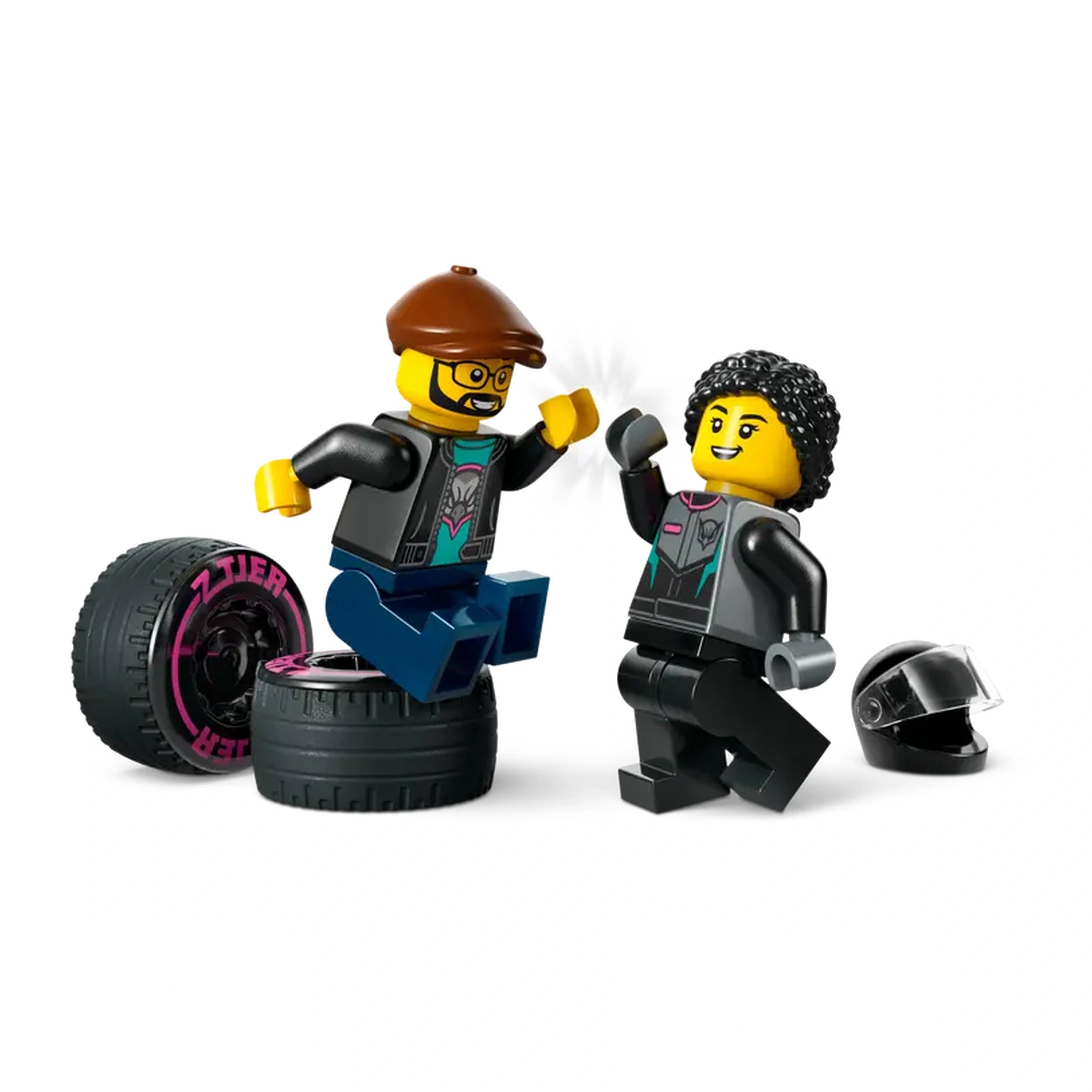 Two LEGO minifigures, a male with glasses and a brown cap and a female with curly hair, celebrate together on black race car tires. They wear matching black outfits with colorful accents and share an excited expression. A black helmet with a clear visor rests nearby.