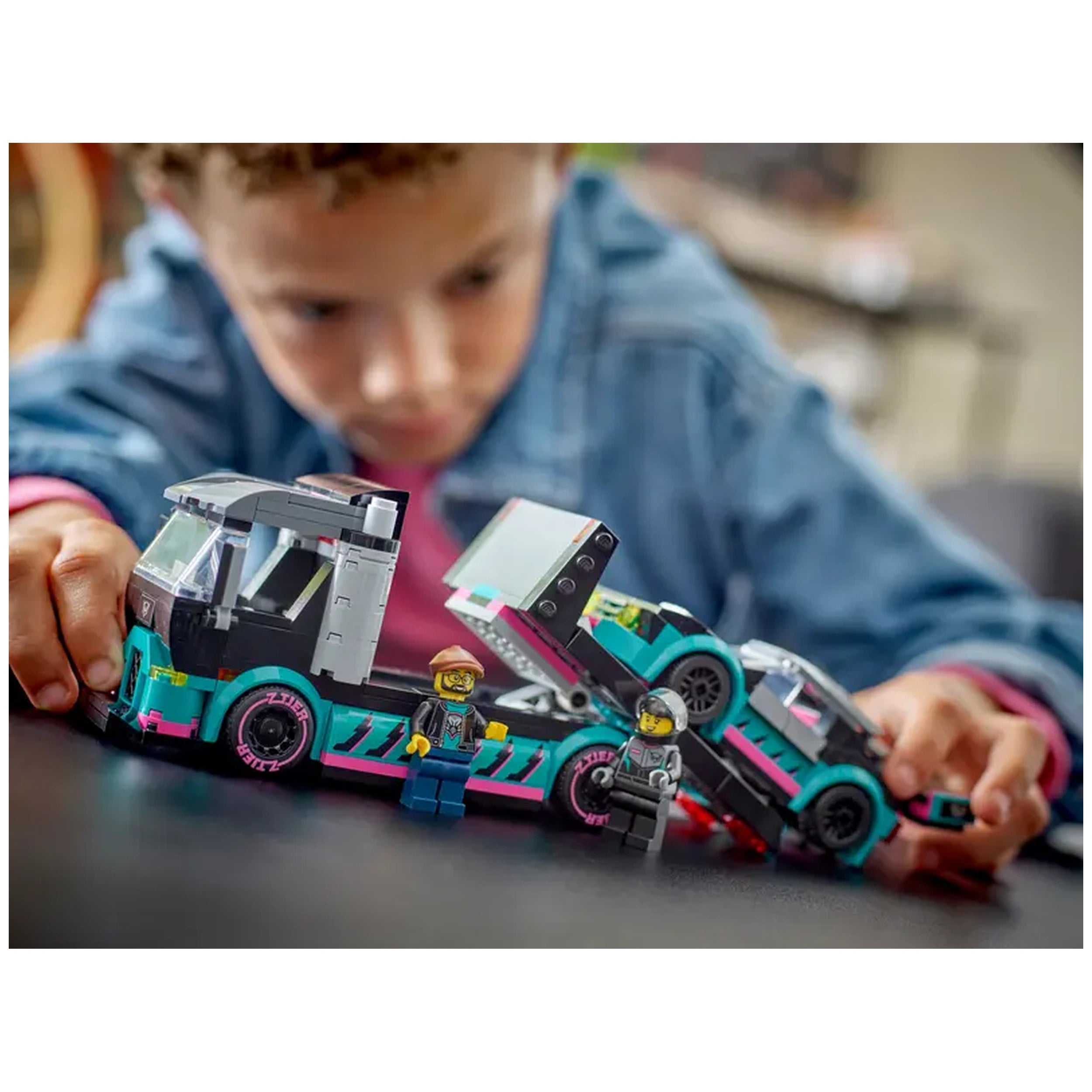 A young child interacts with a LEGO City race car and truck set, focusing on a car carrier truck with vibrant colors. Two minifigures, a truck driver and a race driver, stand beside the vehicles, which feature detailed designs. The scene captures an imaginative moment of play with the truck's loading ramp extended, offering an engaging setup for role-playing scenarios.