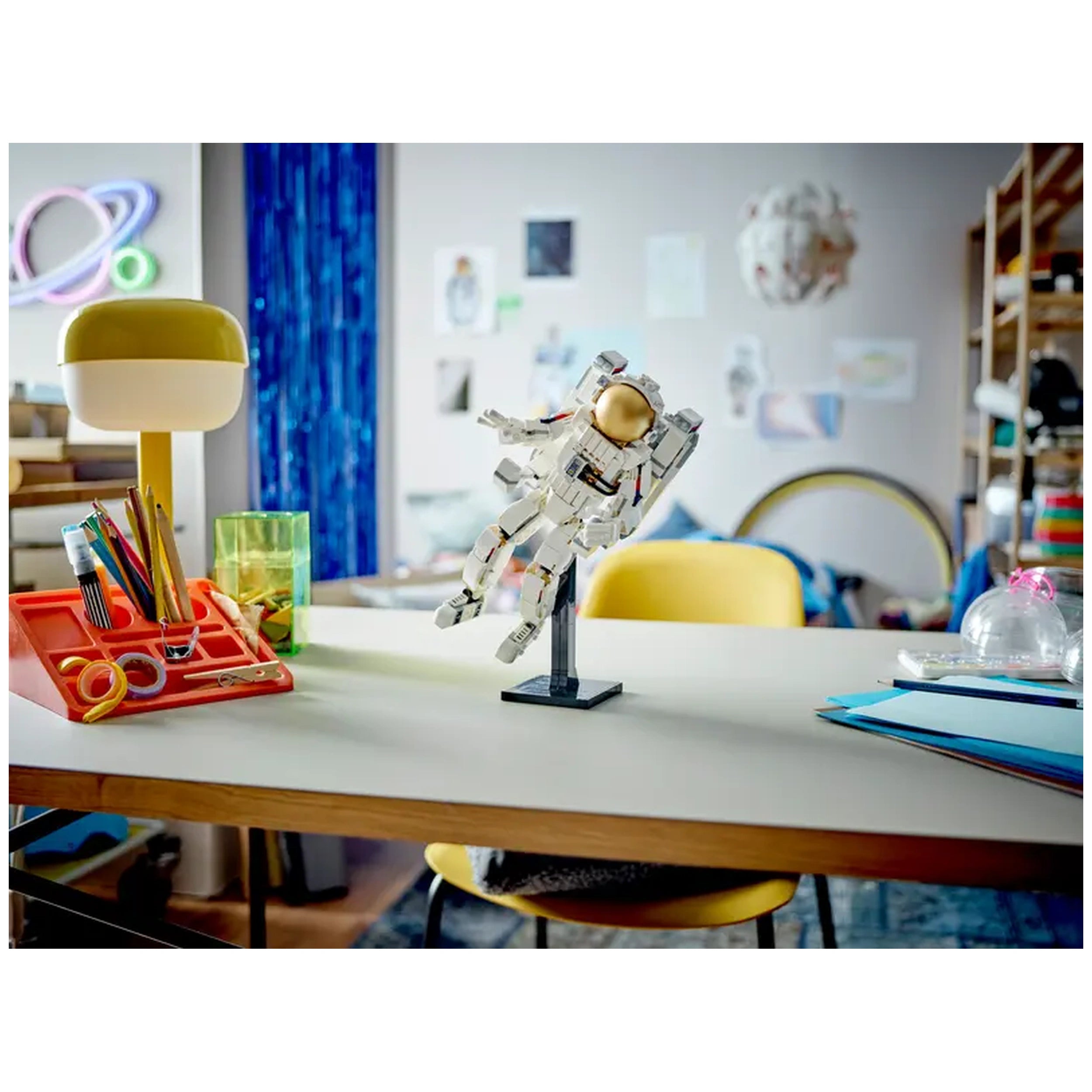LEGO Creator Space Astronaut figure in a playful pose, set against a bright workspace with colorful stationery and a yellow lamp.