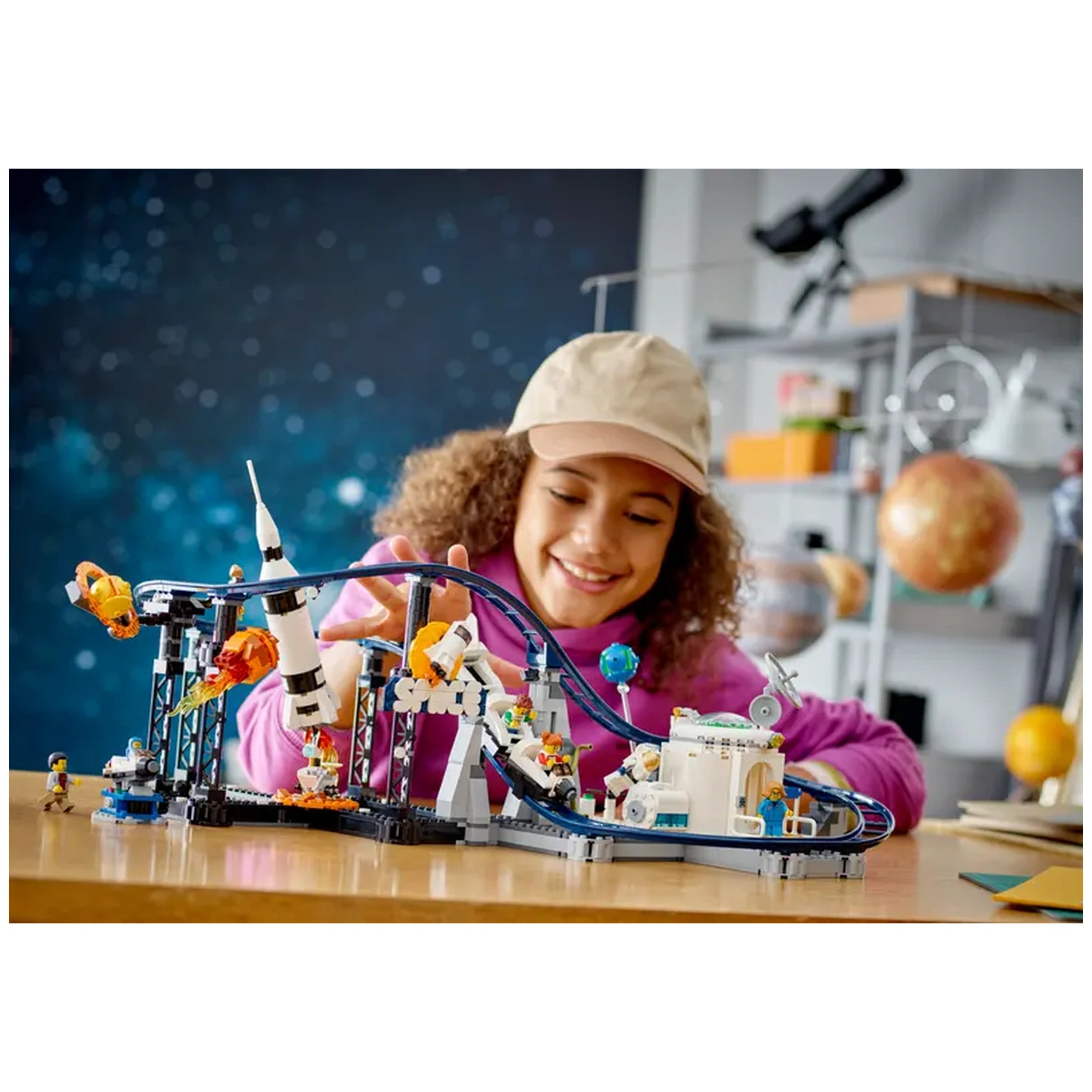 A child smiles while playing with the LEGO® Creator 3in1 Space Roller Coaster set, featuring colorful mini figures, a rocket, and a winding track, all set against a starry background.