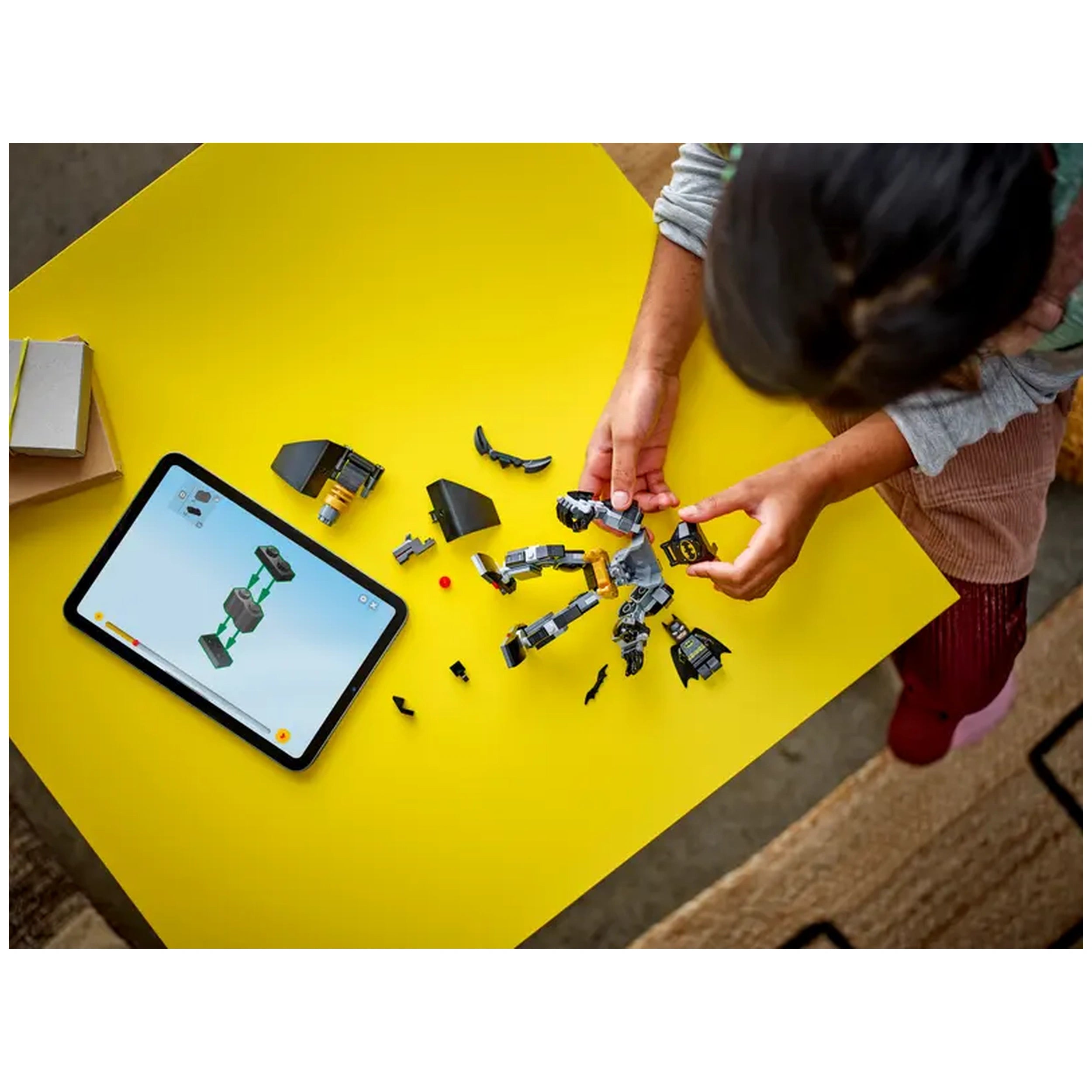 A child's hands assemble the Batman Mech Armor construction set on a bright yellow table, with LEGO pieces scattered around and a tablet displaying building instructions.