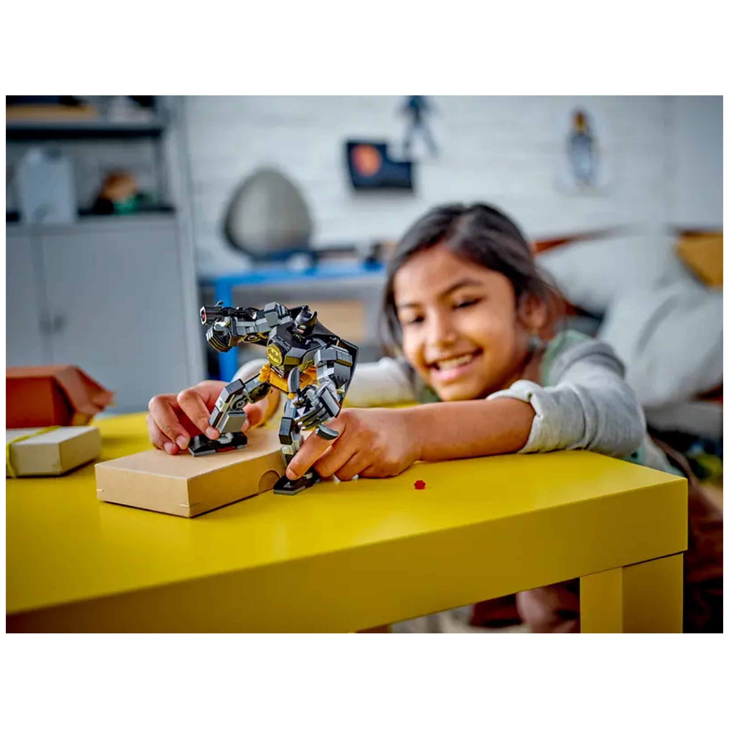 A child joyfully interacts with a Lego Batman Mech Armor on a yellow table, manipulating the mech's pose with excitement.