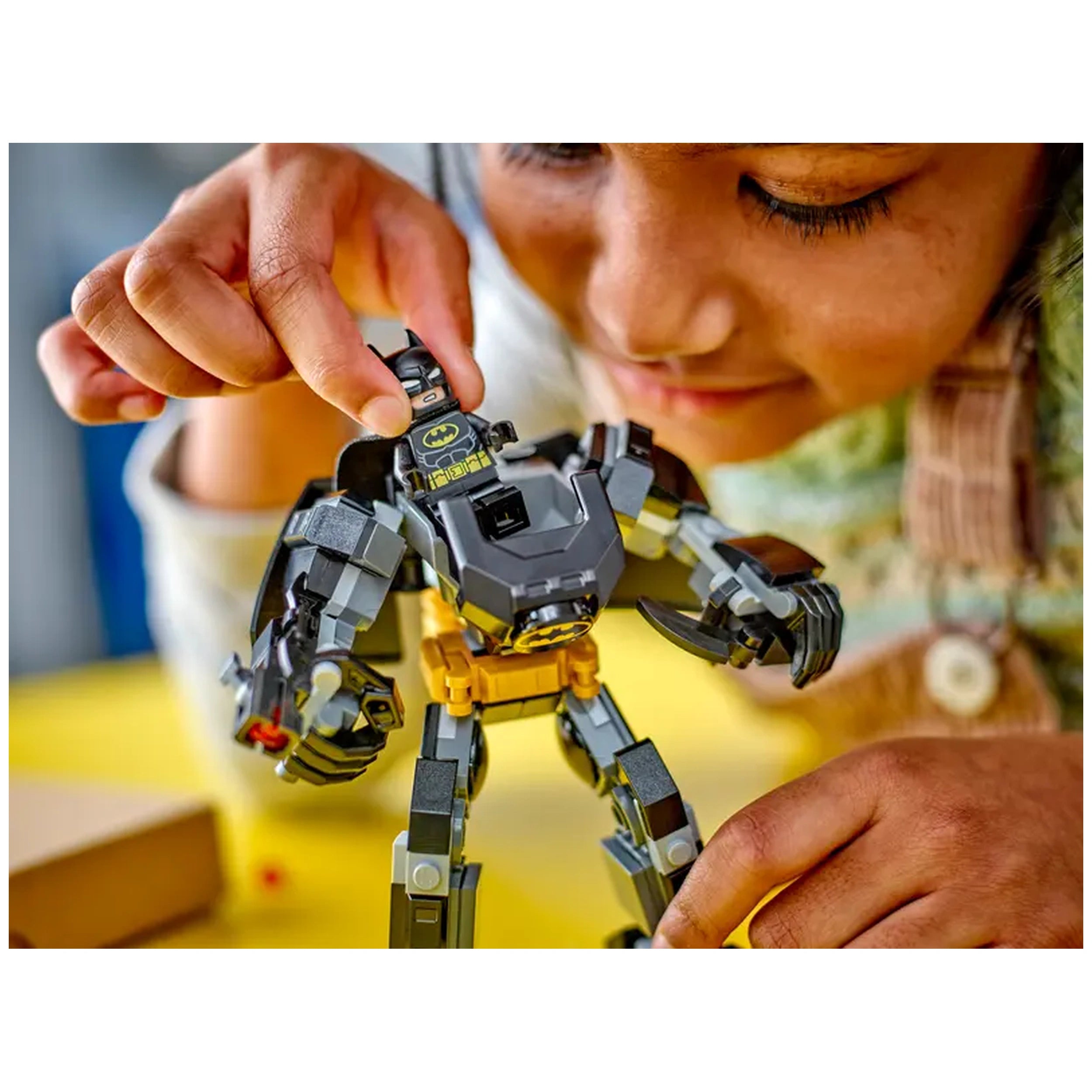 A child plays with a Batman Mech Armor LEGO set, carefully inserting a Batman minifigure into the mech's cockpit, against a bright yellow background.