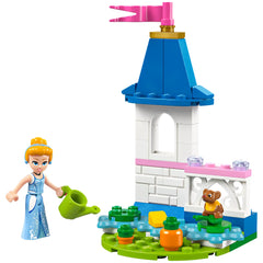 Cinderella, wearing a blue dress, waters vibrant flowers in her mini garden castle with a blue turret and pink flag. A small brown bear sits nearby on green leaves.