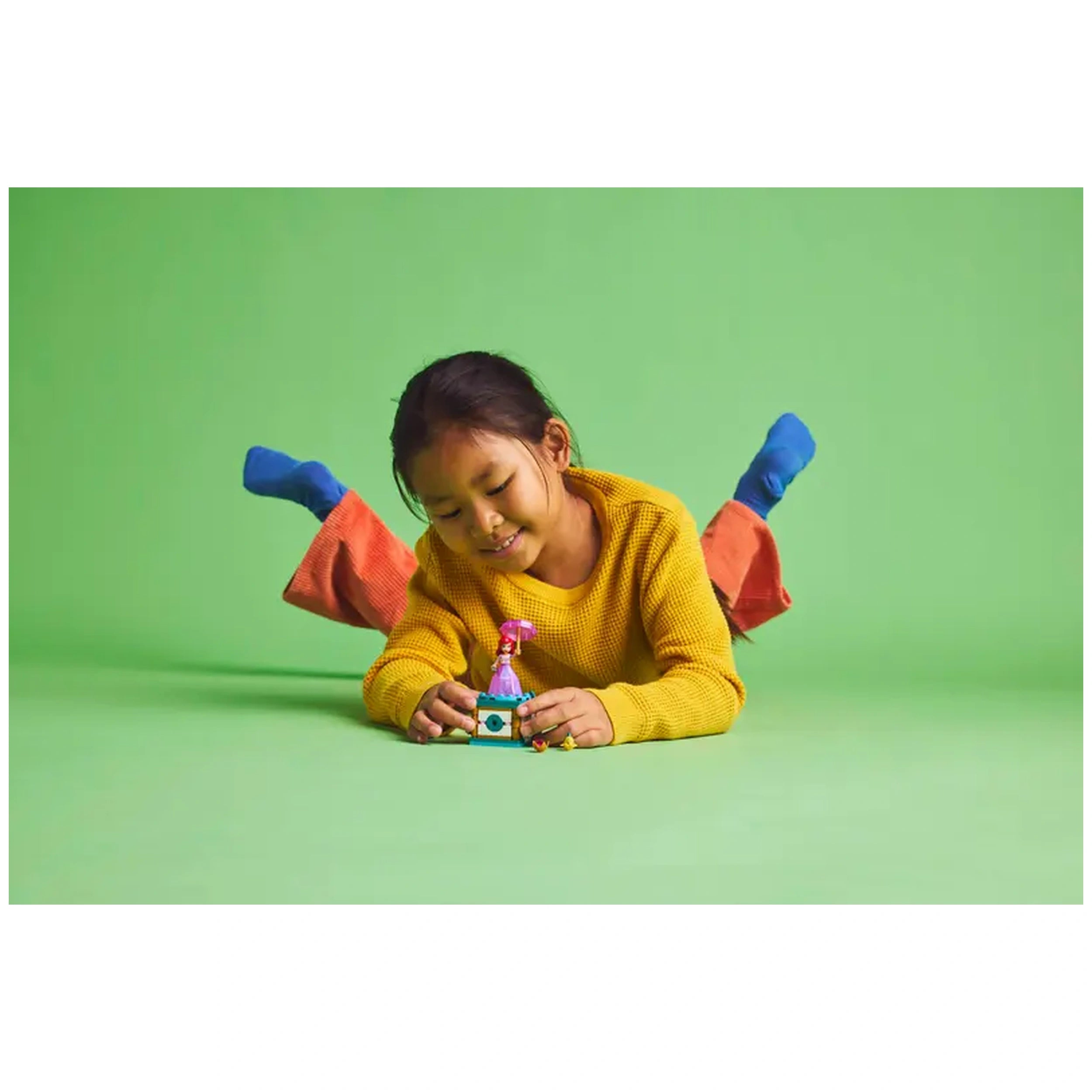 A joyful child wearing a yellow sweater and colorful pants lies on a green background, playing with the LEGO Disney Twirling Ariel construction set.