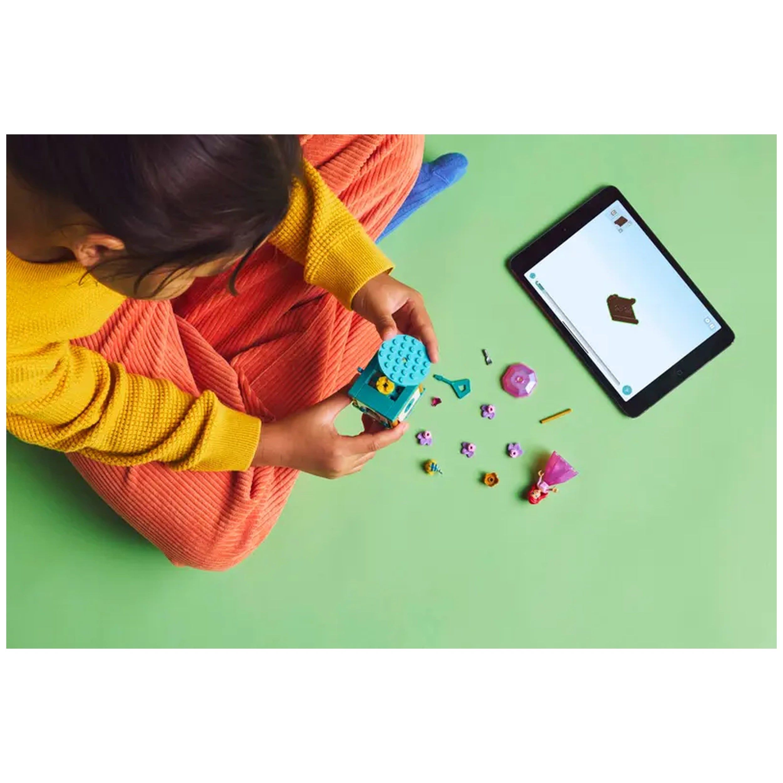 A child in a yellow sweater holds a turquoise LEGO® box while sitting on a green floor, surrounded by colorful accessories, including flowers and a mini doll. An iPad displaying a building guide is positioned nearby.