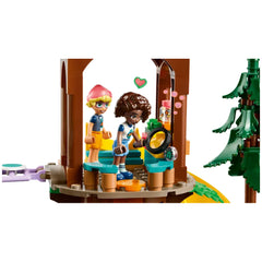 Two mini-dolls, one with glasses and a pink hat, explore a tree house with colorful seating. They examine nature using a magnifying glass, surrounded by playful accessories. A green tree stands nearby, enhancing the outdoor adventure theme.