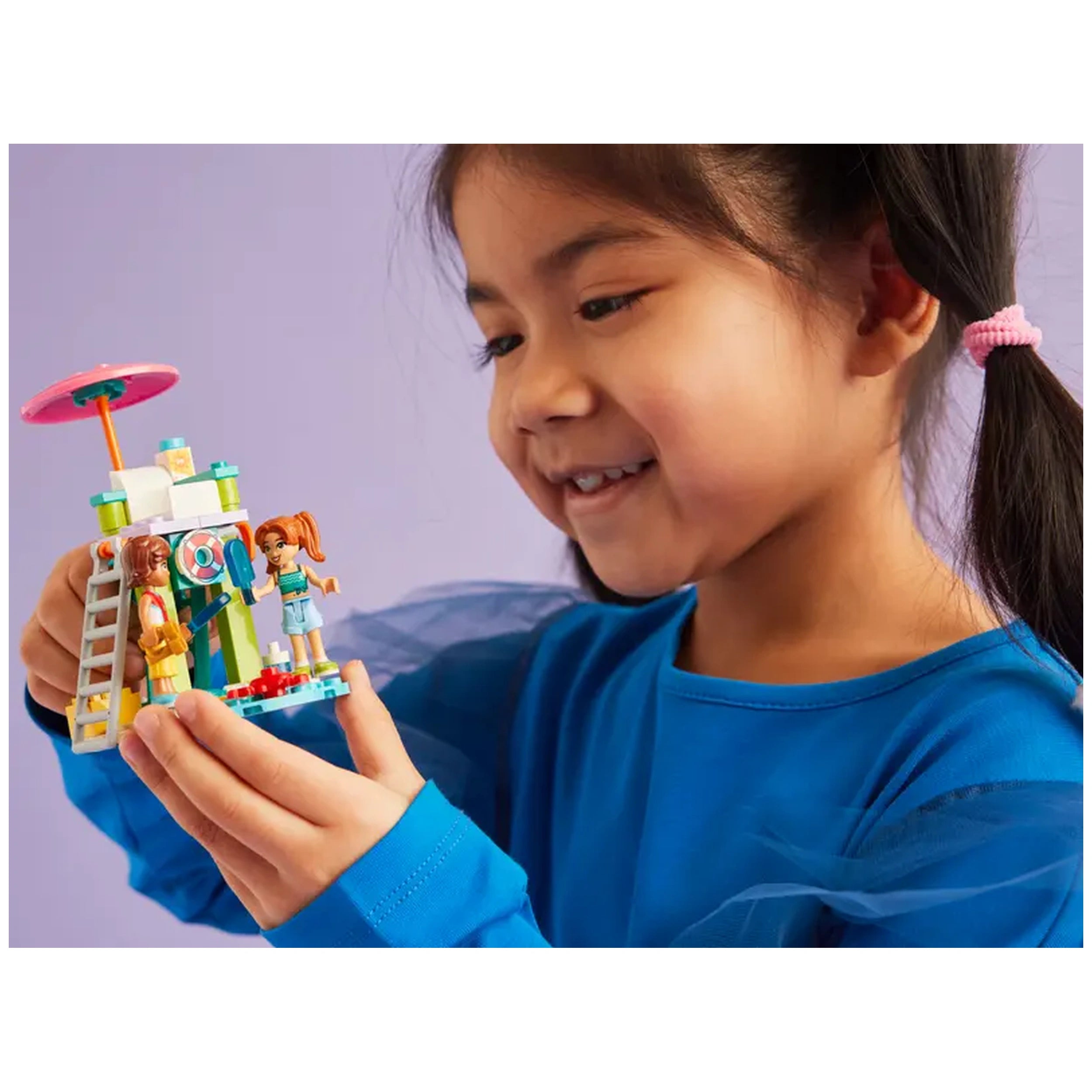 A child in a blue shirt smiles as she holds the LEGO Friends Beach Water Scooter construction set, featuring two mini-dolls and colorful accessories.